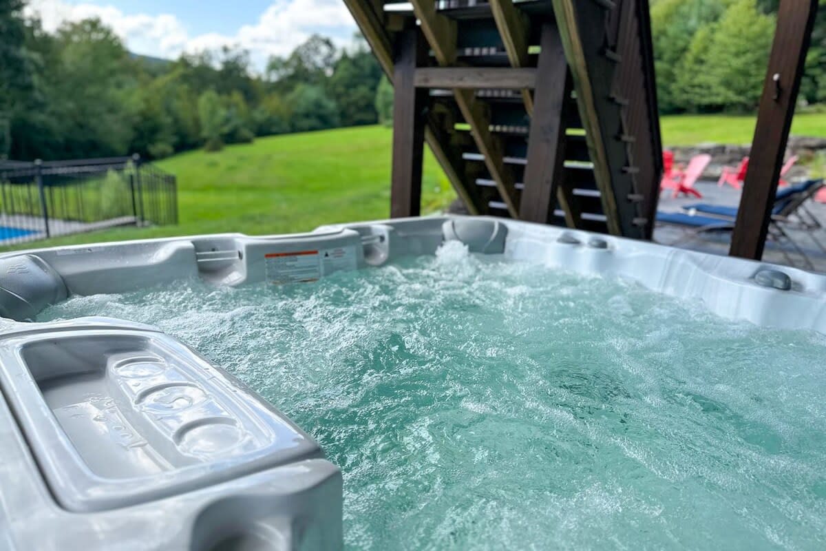 A bubbling hot tub with light gray exterior is positioned on a deck, offering a view of a lush green hillside and a distant swimming pool area, under a bright sky.