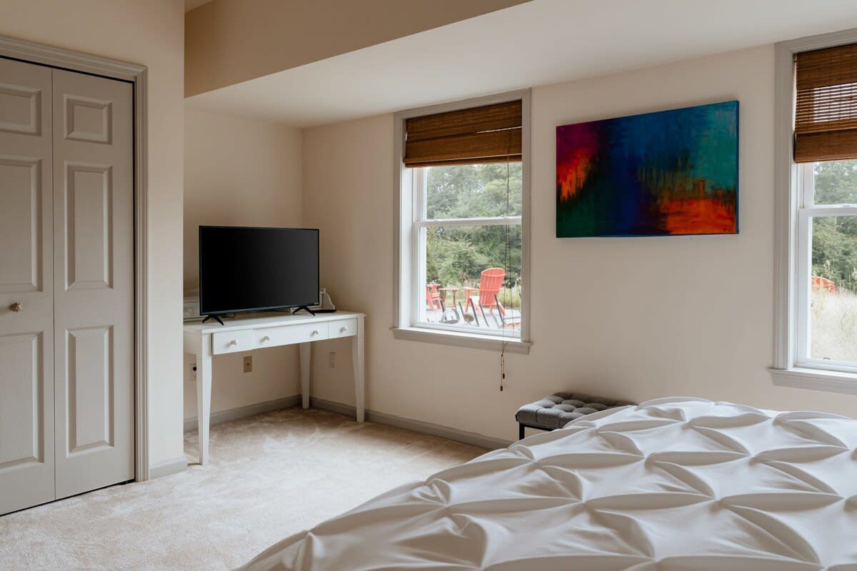 A bright bedroom with a white bed, a dark flat-screen TV on a white desk, and a window overlooking an outdoor patio. A colorful abstract painting hangs on the light-colored wall.