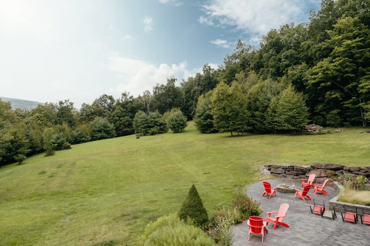 A spacious, gently sloping green lawn is surrounded by dense trees and features a paved patio area with several red Adirondack chairs and a fire pit, offering expansive views of the natural landscape.