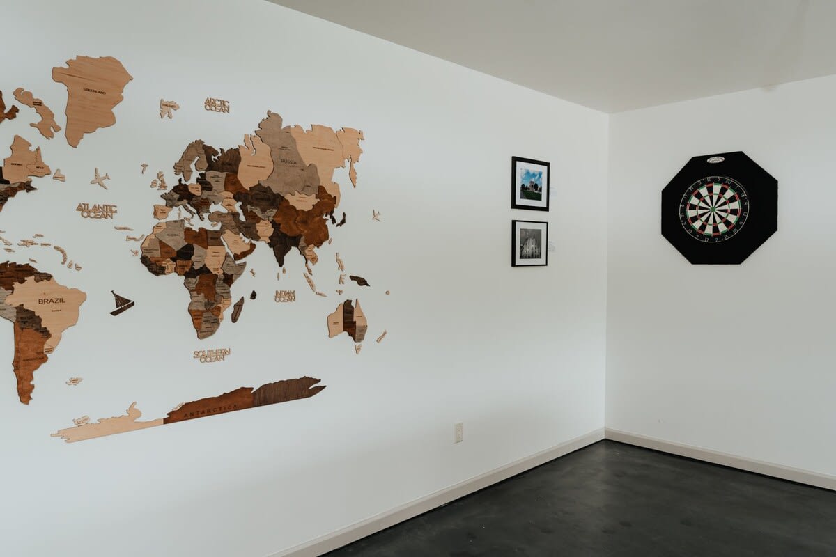A white-walled room featuring a large wooden world map art piece and a dartboard, with dark flooring, suggesting a dedicated recreation space.