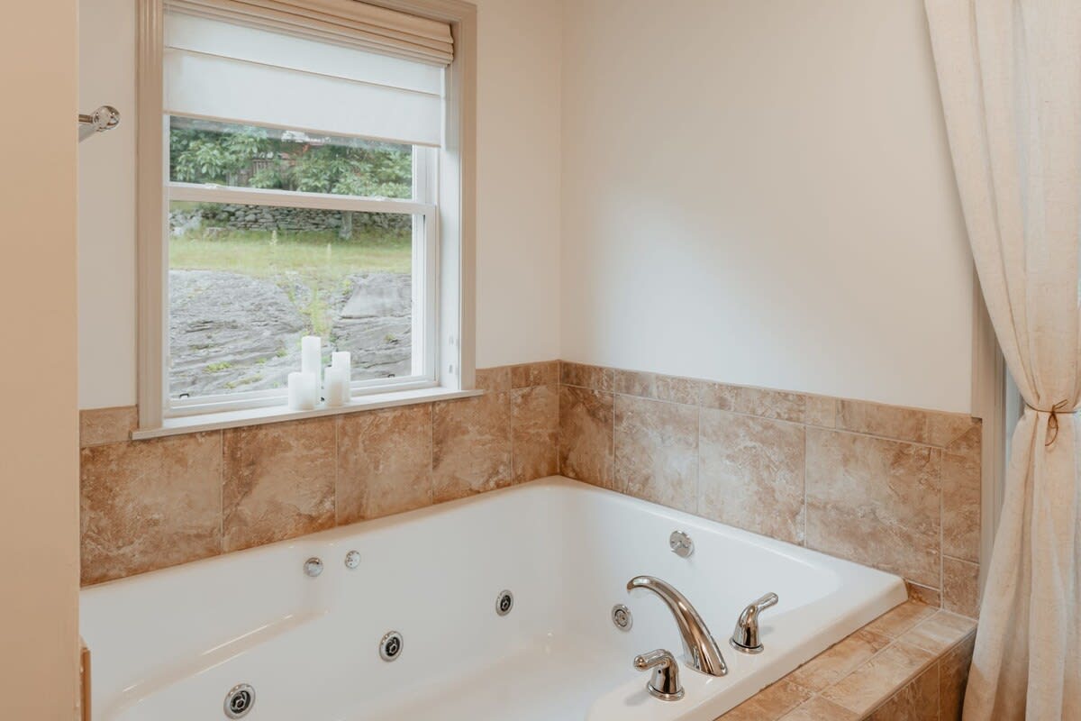 A close-up of the jetted bathtub with a light brown tiled surround, positioned beneath a window with a beige Roman blind, showcasing decorative candles on the windowsill.
