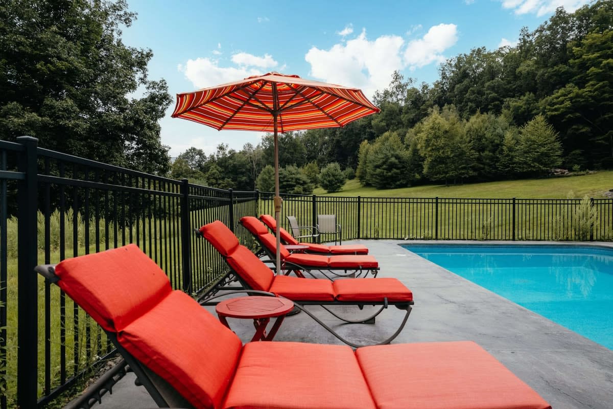 A rectangular swimming pool with bright blue water is bordered by a dark fence and a concrete deck, featuring several orange lounge chairs and a striped umbrella, with a green hillside in the background.