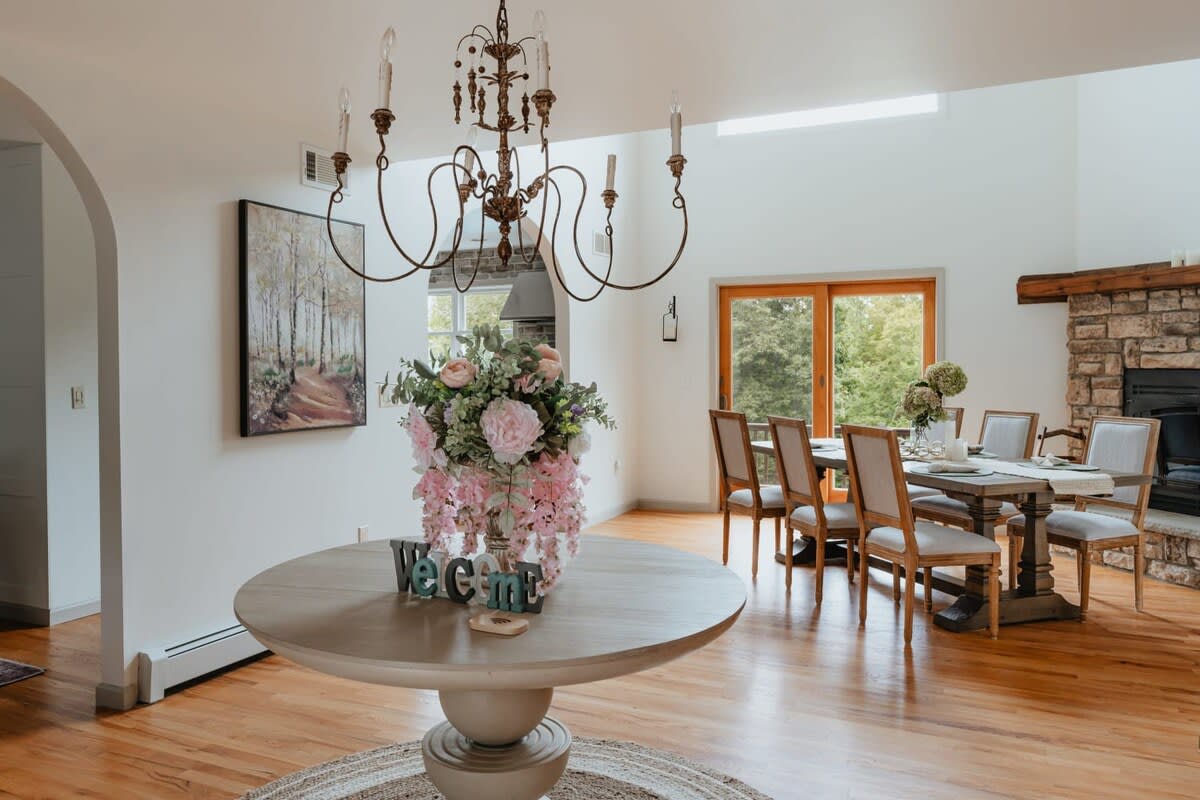 A spacious foyer area with a round wooden table adorned with a large floral centerpiece, under a rustic chandelier. In the background, a dining area with a wooden table and chairs is visible through an archway, with large glass doors leading outside.