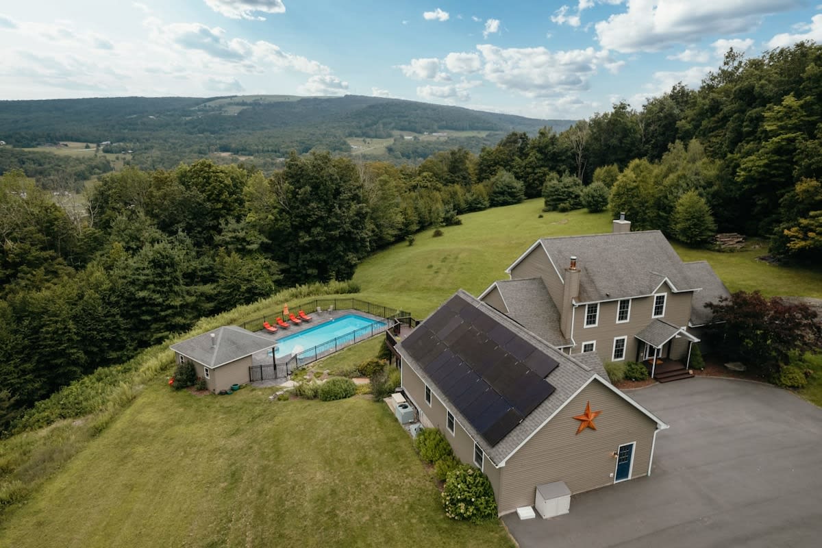 An aerial view of a large house with a gray roof and solar panels, situated on a sprawling green hillside with a fenced-in swimming pool and pool house, surrounded by lush trees and distant mountains.