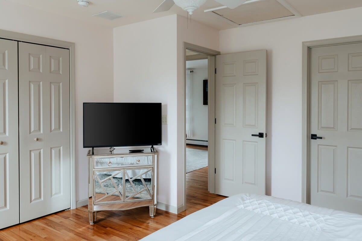 A view of the bedroom showing a flat-screen TV on a mirrored dresser, two light gray bi-fold closet doors, and an open doorway leading to another room.