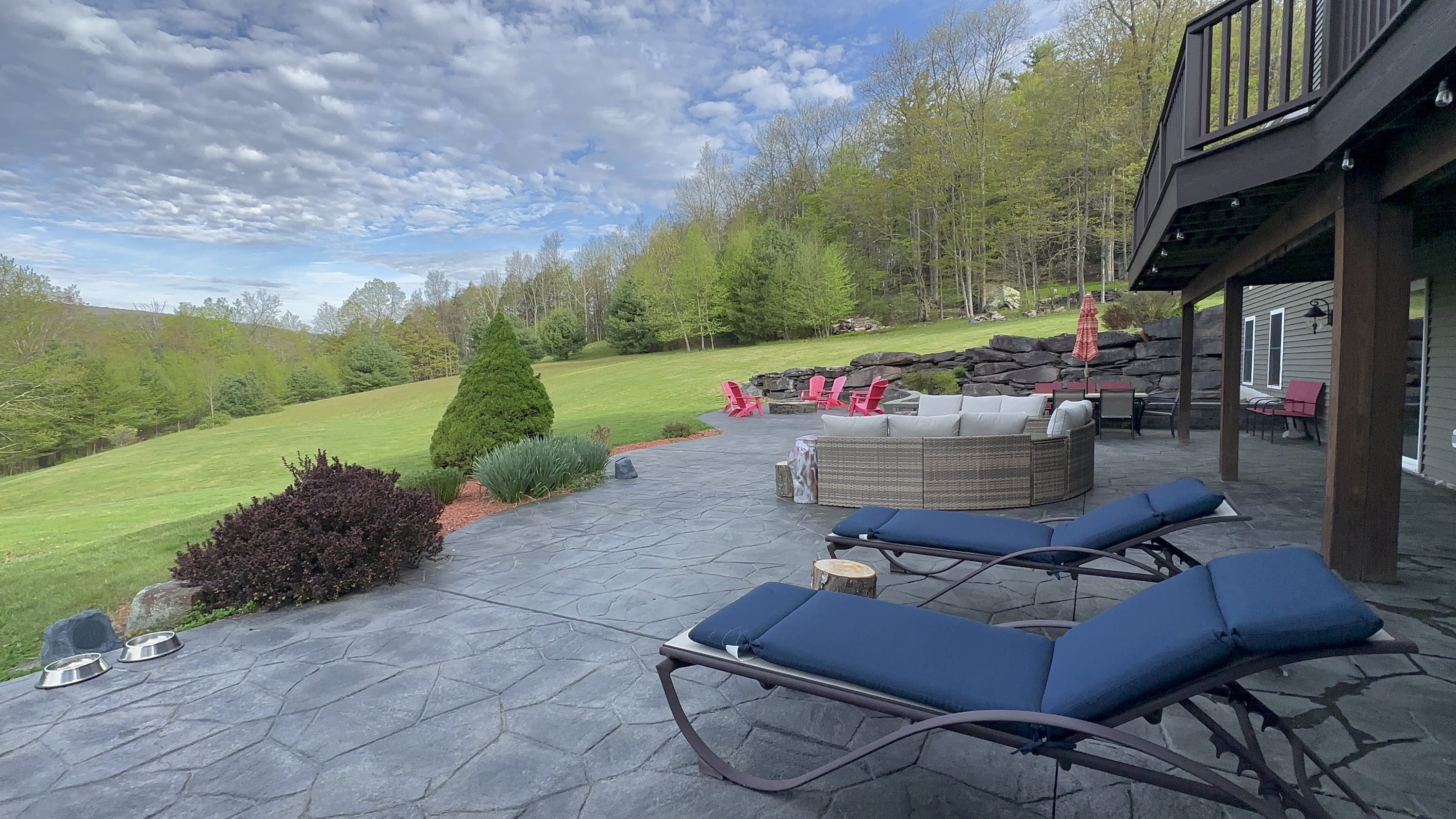 A wide view of the stamped concrete patio featuring two blue lounge chairs and a wicker sectional sofa. The expansive green lawn slopes up to a wooded area, with a fire pit and red chairs in the distance.