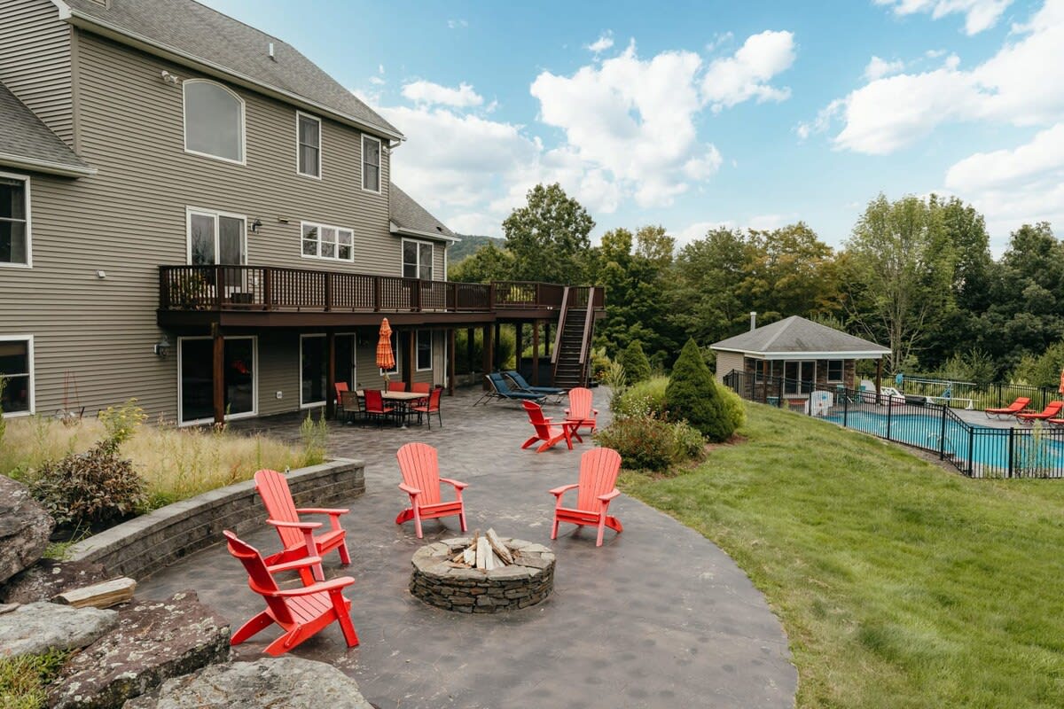 The back exterior of a multi-story house with light gray siding, featuring a large deck, a stamped concrete patio with a stone fire pit and red Adirondack chairs, and a fenced swimming pool area.