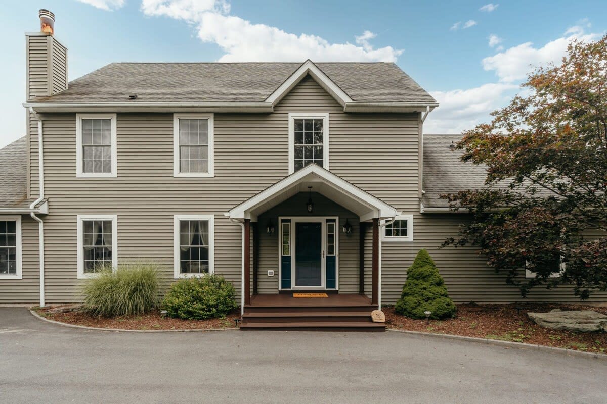 The front facade of a two-story house features light green horizontal siding, white-trimmed windows, and a dark gray shingled roof with a prominent chimney. A welcoming covered porch with dark wood pillars leads to a dark blue front door, flanked by manicured shrubs and a small evergreen.