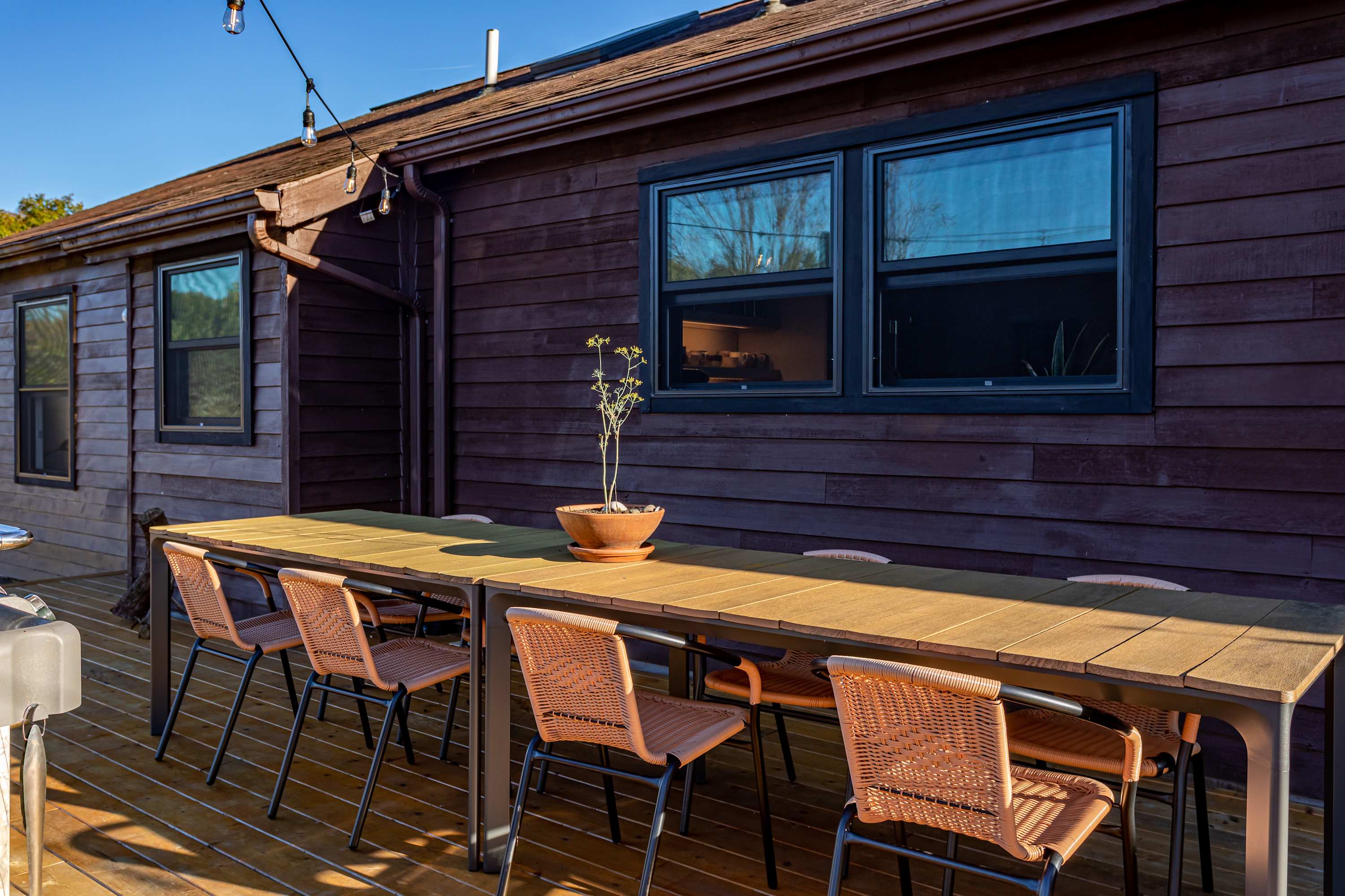 A spacious outdoor dining area on a wooden deck features a long rectangular table with a light wood top and dark metal frame, surrounded by eight woven-back chairs. String lights hang overhead, and a potted plant serves as a centerpiece.