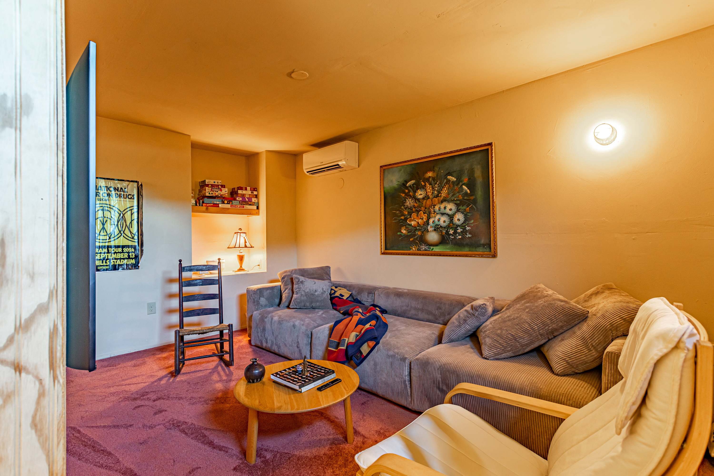 A comfortable living space features a large grey sectional sofa with corduroy upholstery and an accent blanket, facing a light wood coffee table on a plush reddish-brown carpet. A vintage rocking chair and a built-in shelf with a lamp add character.
