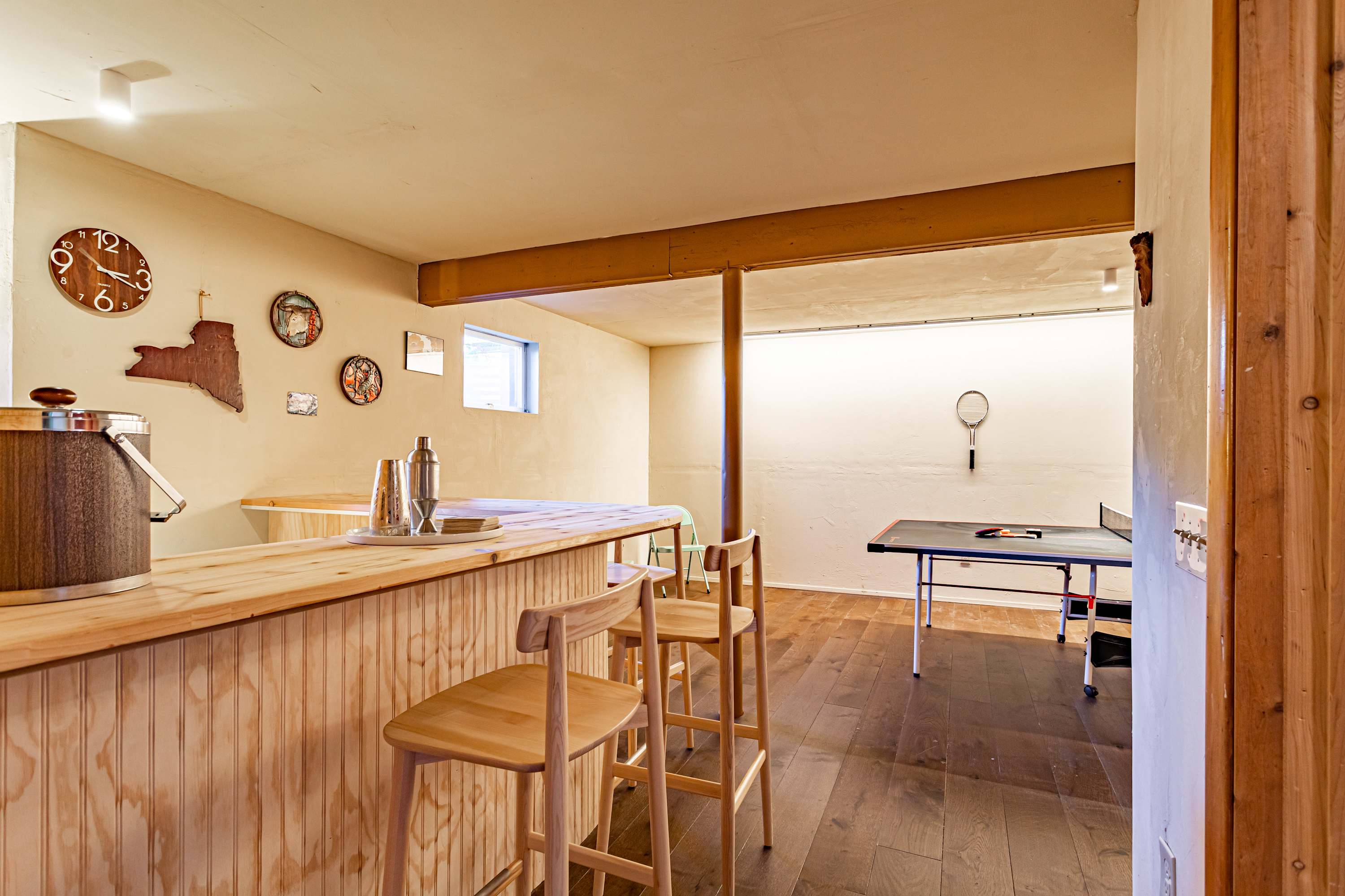 A basement game room featuring a wooden bar with bar stools and a ping-pong table, set against light-colored walls and dark wood floors, offering a casual entertainment space.