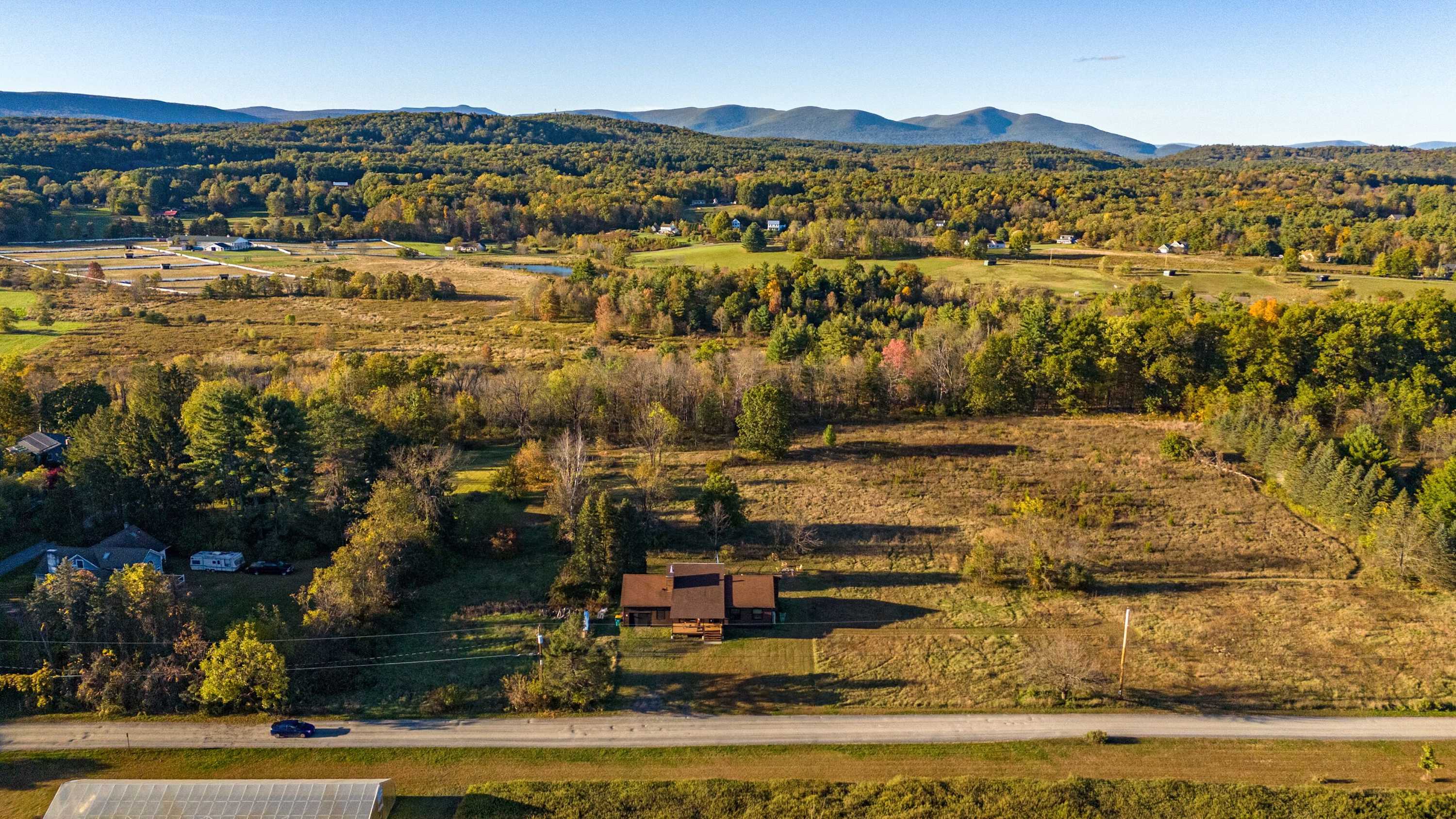 An aerial view showcases the property nestled amidst expansive fields and lush trees, with a road running in the foreground. Distant mountains form a scenic backdrop under a clear blue sky, highlighting the rural setting.