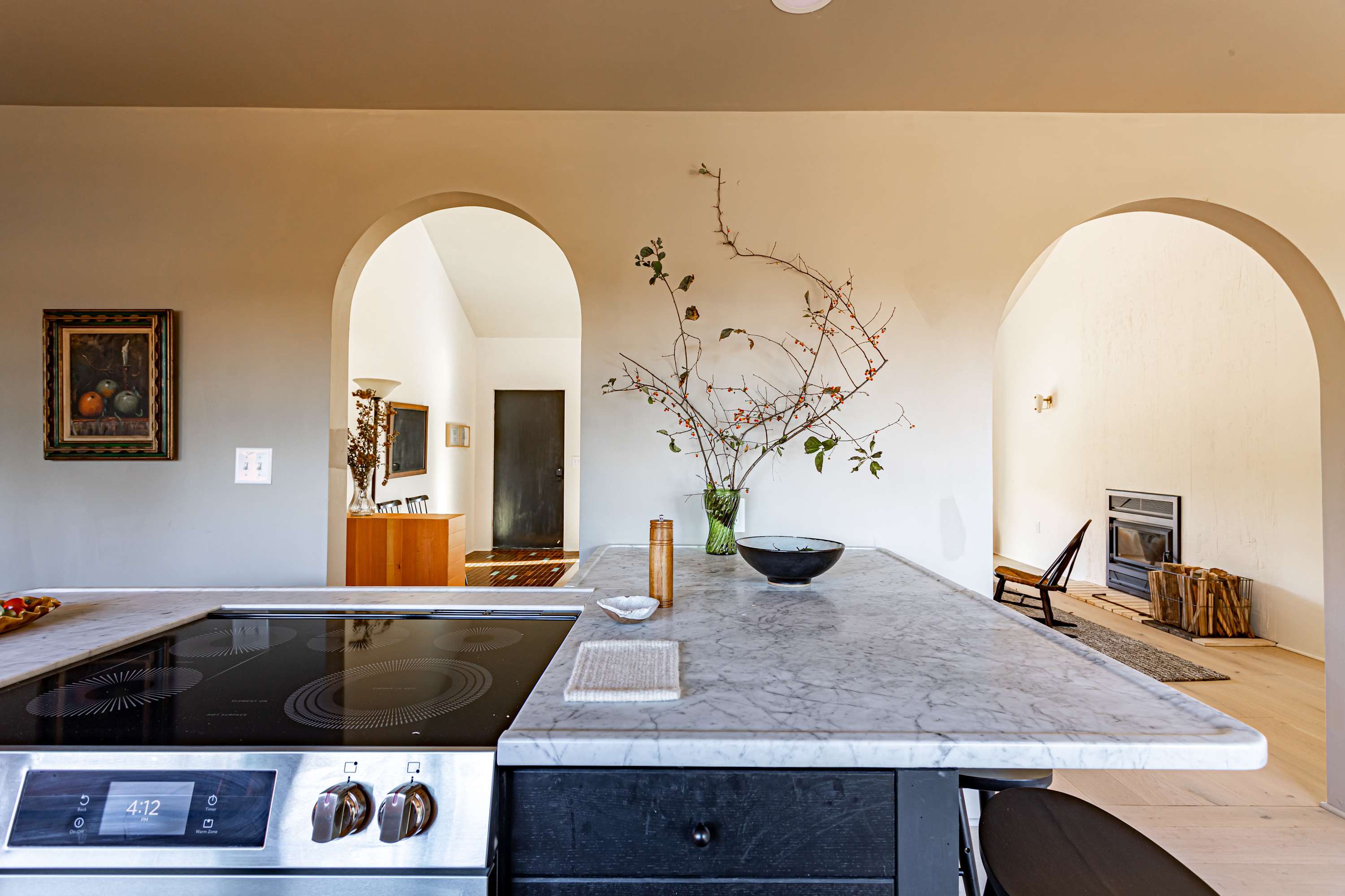 Kitchen island with a marble countertop and a built-in electric cooktop, offering a view into the living area through arched openings. A vase with branches and a black bowl decorate the island.