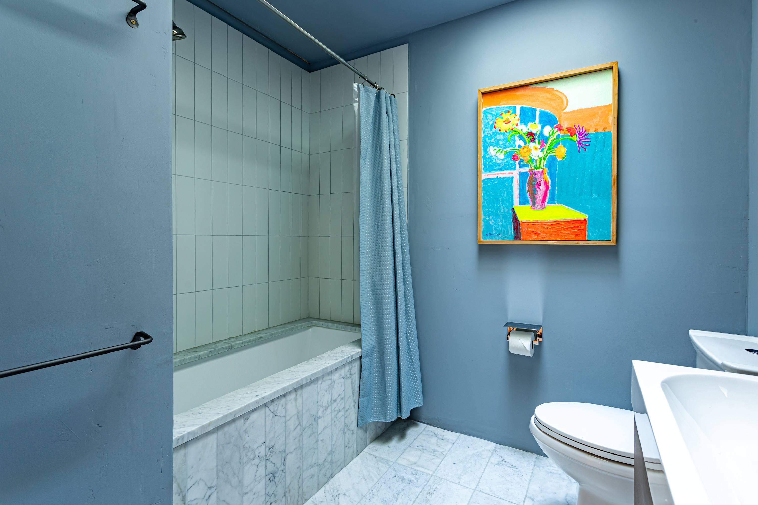 A vibrant blue bathroom featuring a white bathtub with subway tile surround, a blue shower curtain, a toilet, and a white vanity with a sink. A colorful abstract painting adds a pop of color to the blue walls.