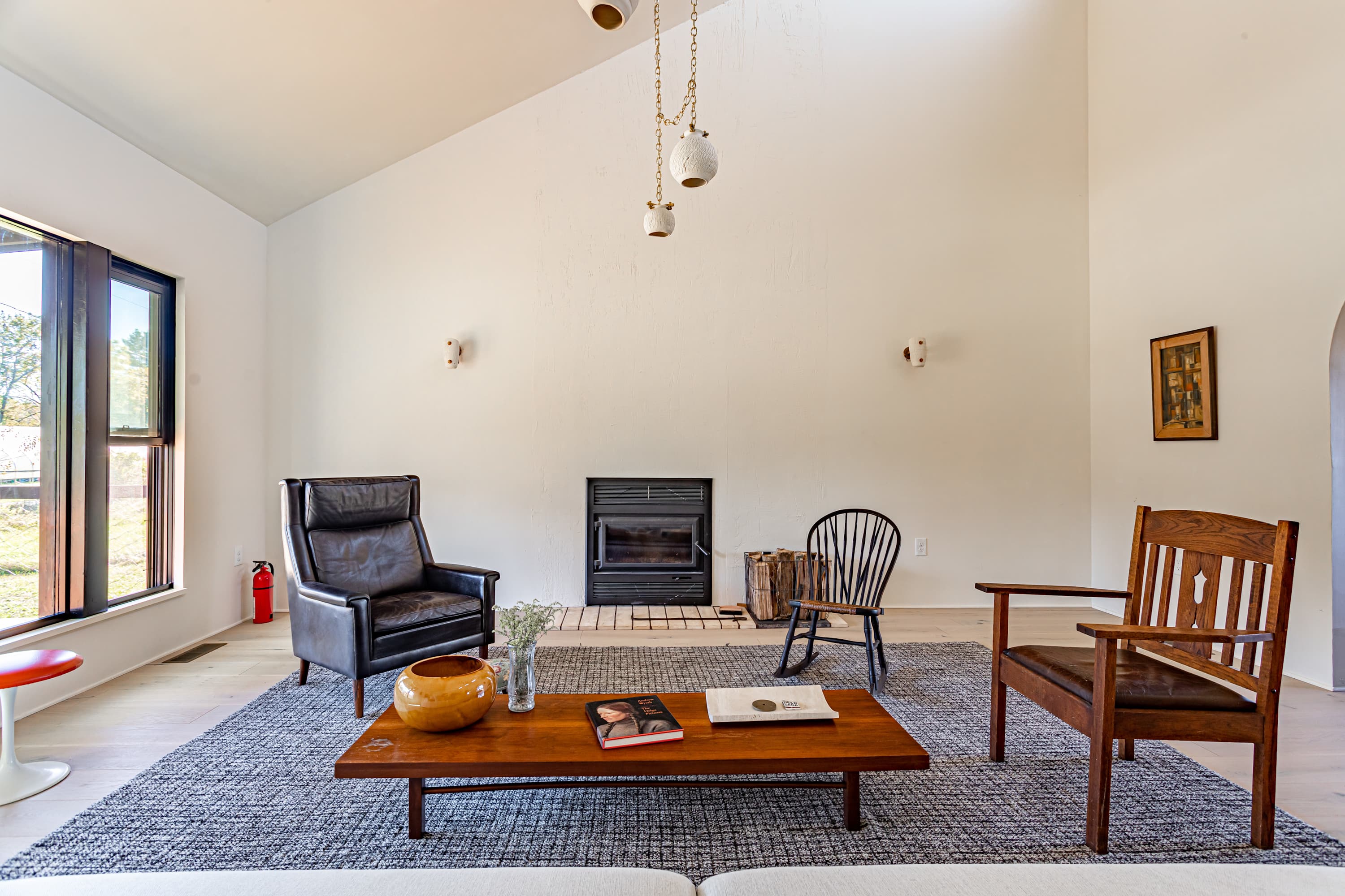 Cozy living room with a black leather armchair, a wooden coffee table on a grey rug, and a corner fireplace. A black rocking chair and a wooden armchair complete the seating arrangement under a vaulted ceiling.