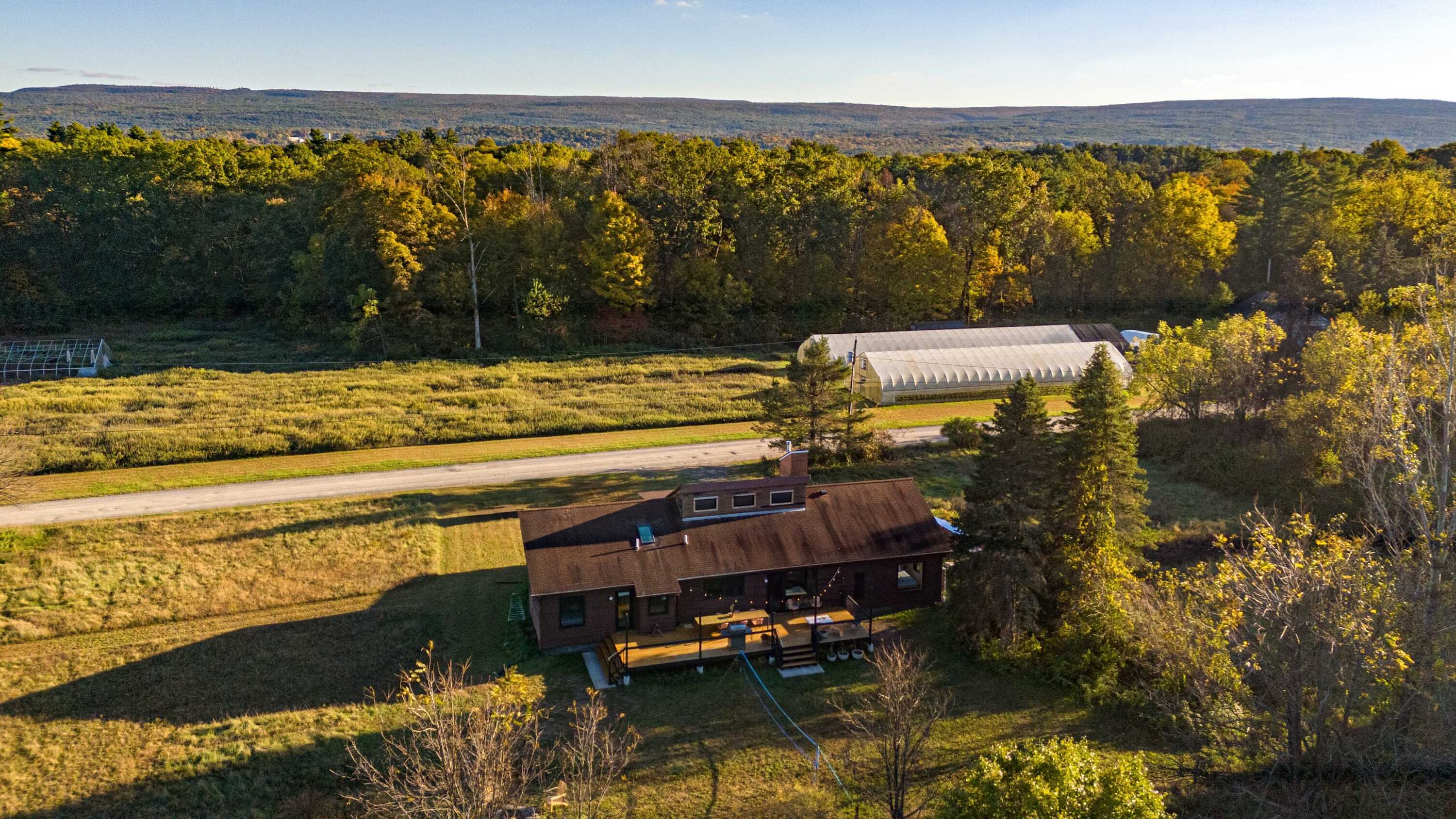 An aerial view showcases the brown-sided house with its large wooden deck, nestled amidst a vast landscape of autumn trees and open fields. A road runs alongside the property, with greenhouses visible in the distance.