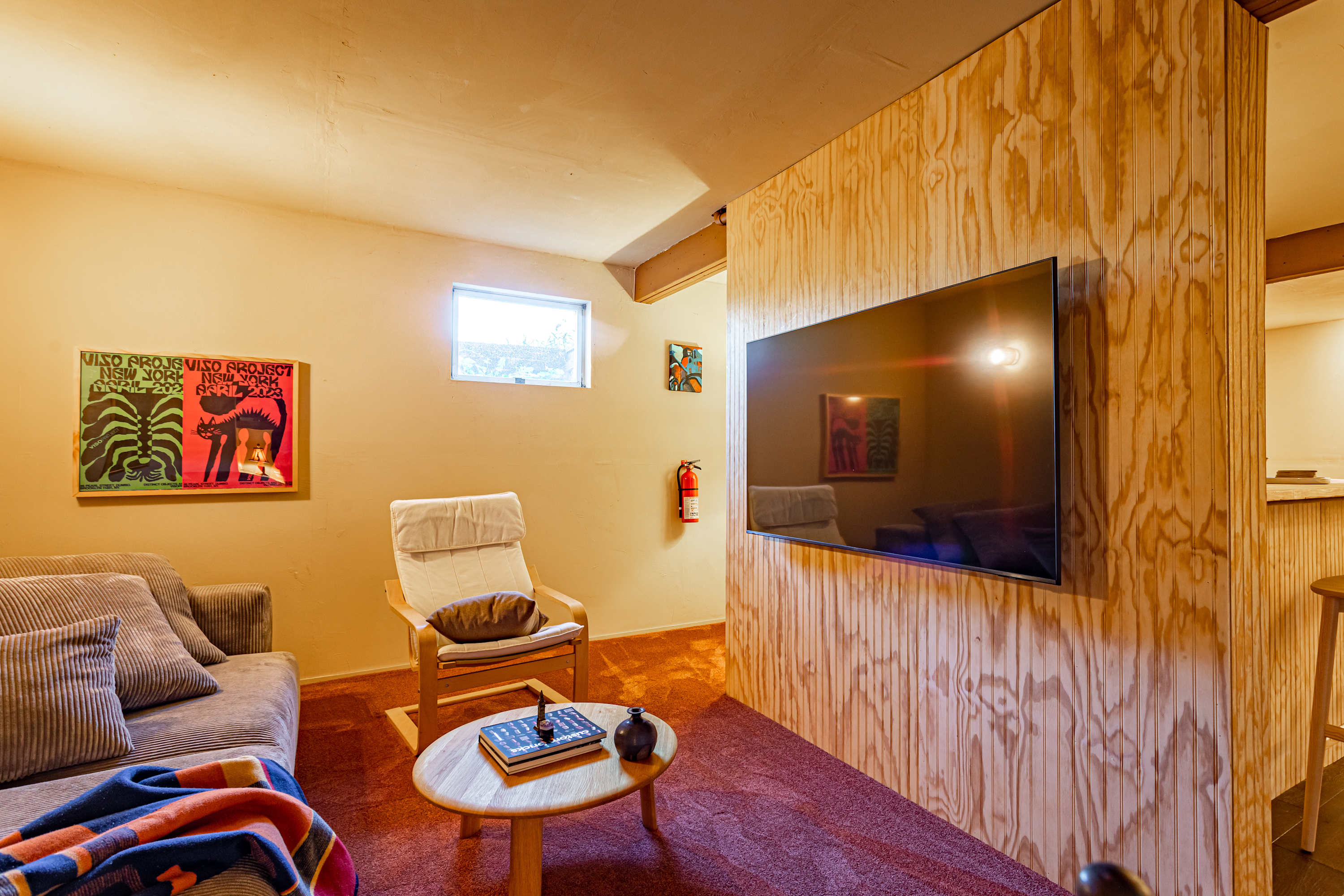 A modern flat-screen TV is mounted on a textured plywood wall, separating a seating area with a grey corduroy sofa and a light wood armchair from the bar area. A small round coffee table sits on a reddish-brown carpet.