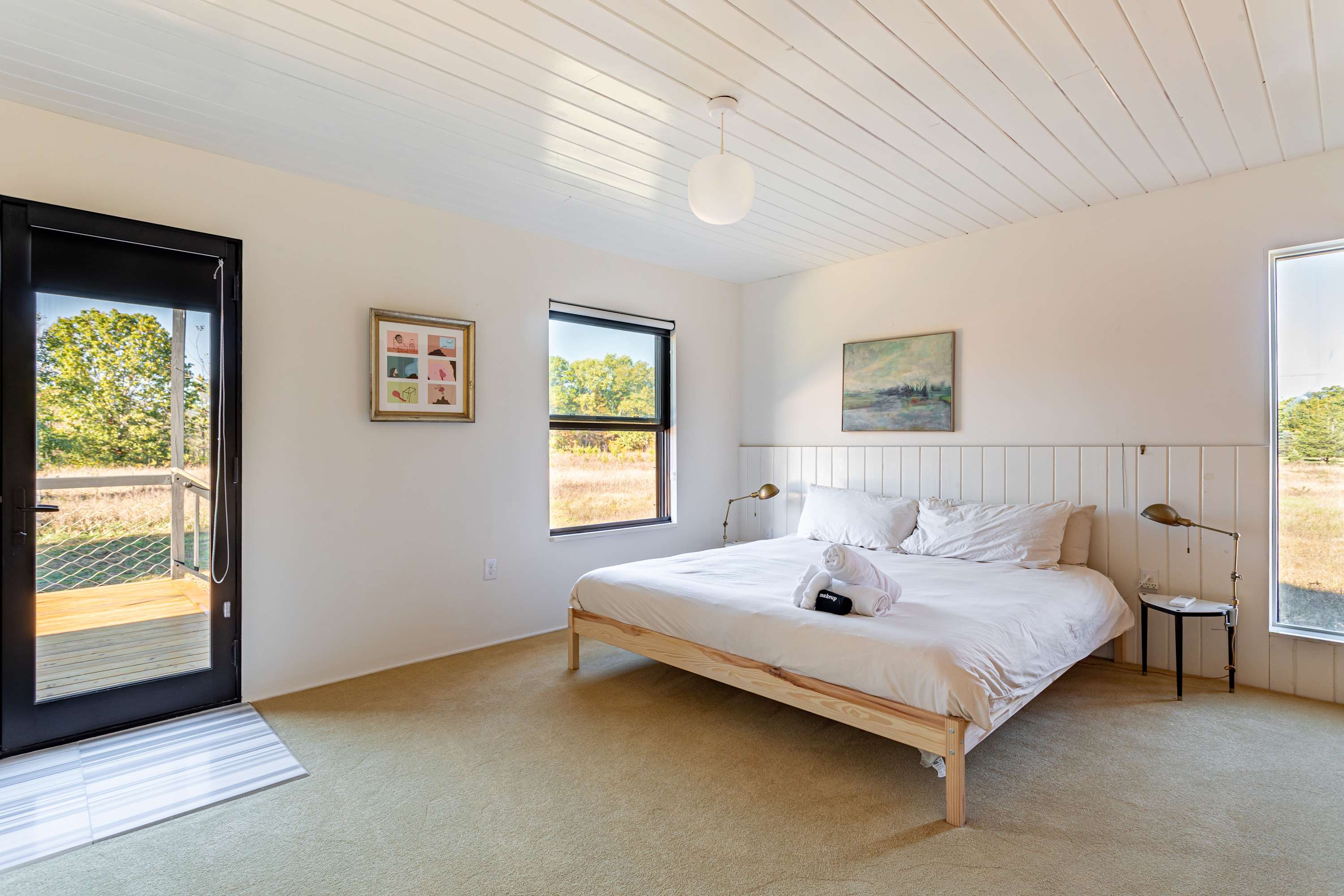 A bright bedroom featuring a queen bed with white linens and a slatted headboard, with a glass door leading to an outdoor deck. Two windows offer views of the surrounding landscape.