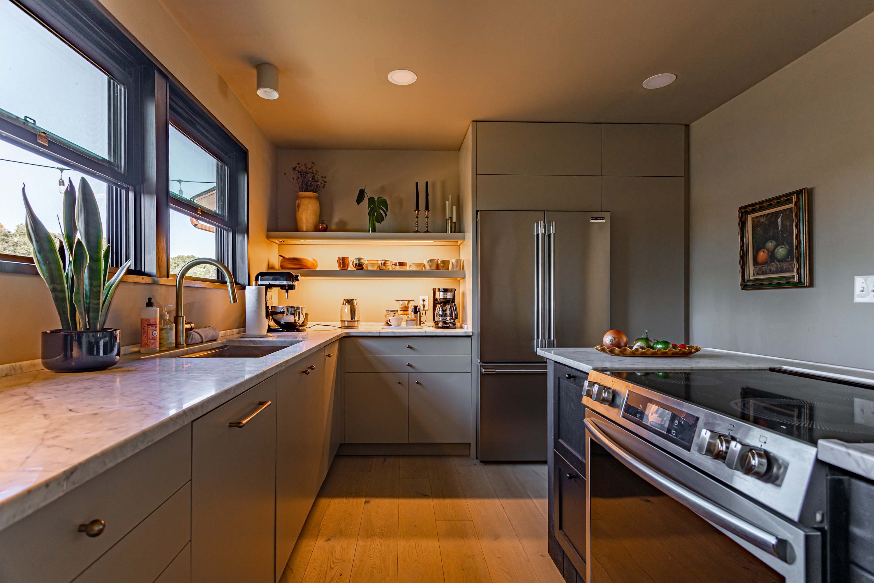 Modern kitchen with light grey cabinetry, white marble countertops, and open shelving with under-cabinet lighting. Stainless steel appliances, including a refrigerator and a range, are visible, along with a sink and various kitchen tools.