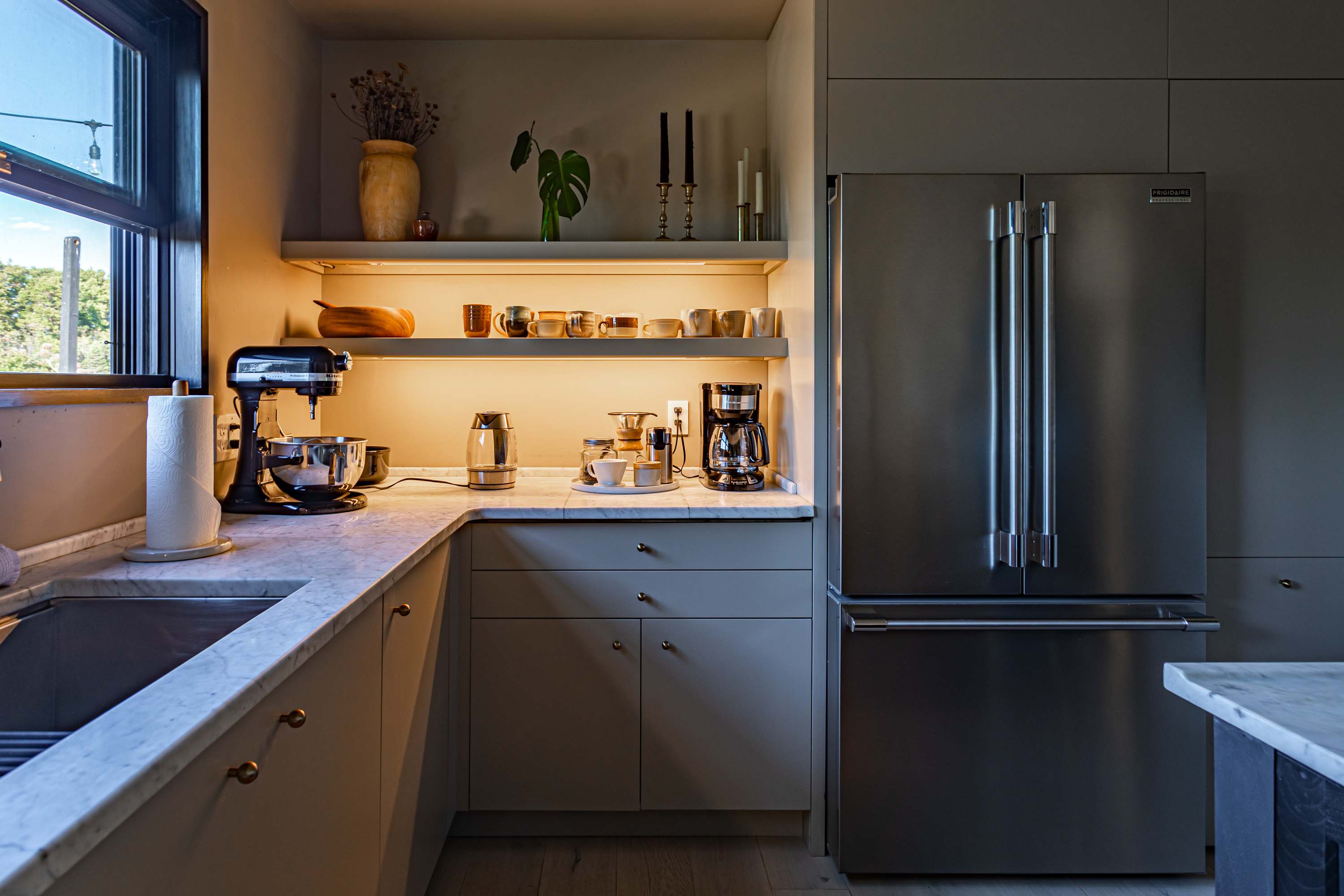 A detailed view of the modern kitchen showing light gray cabinets, white countertops, a stainless steel refrigerator, and open shelving with under-cabinet lighting displaying various kitchenware.