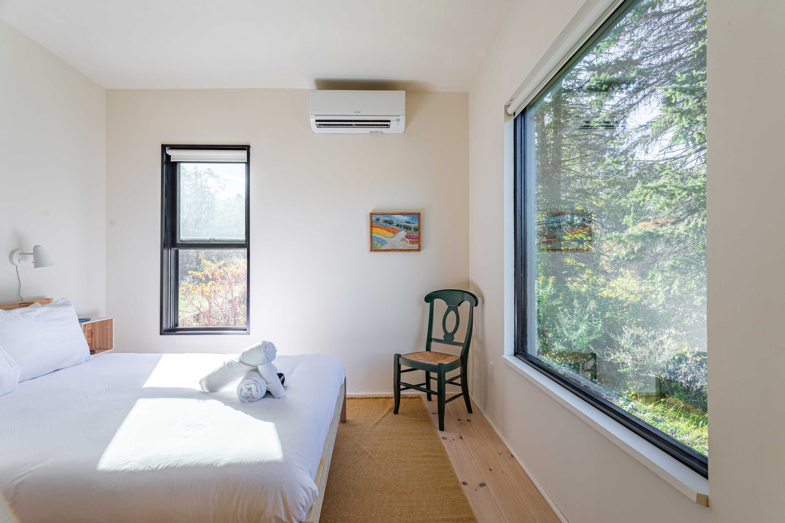 A serene bedroom showcasing a queen bed with white linens and a light wood frame, positioned next to a large window offering expansive views of lush greenery. A dark green wooden chair sits nearby.