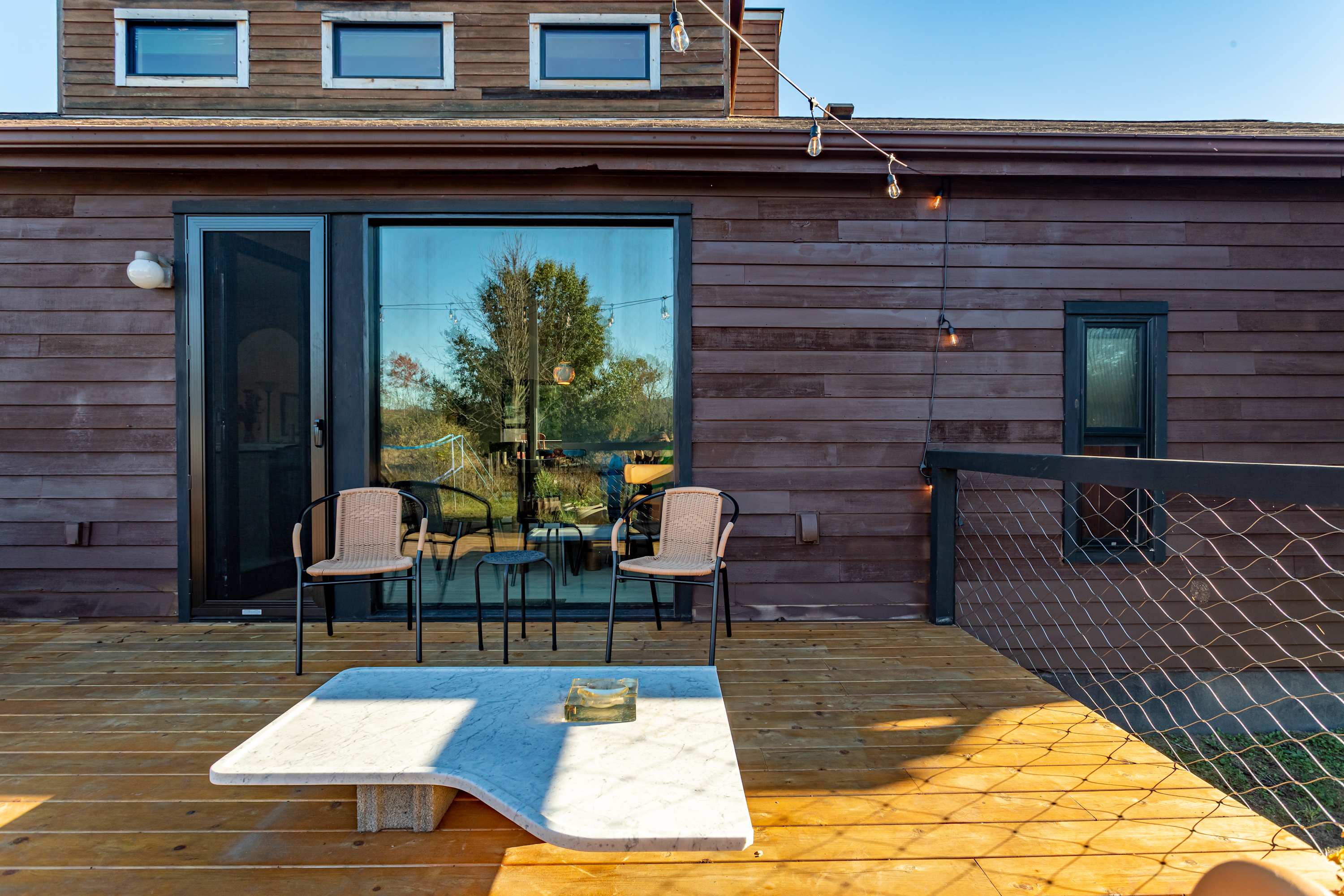 A close-up of the wooden deck features a white marble-top coffee table and two wicker chairs, set against the dark brown siding of the house. String lights are strung overhead, and a large sliding glass door reflects the outdoor scenery.