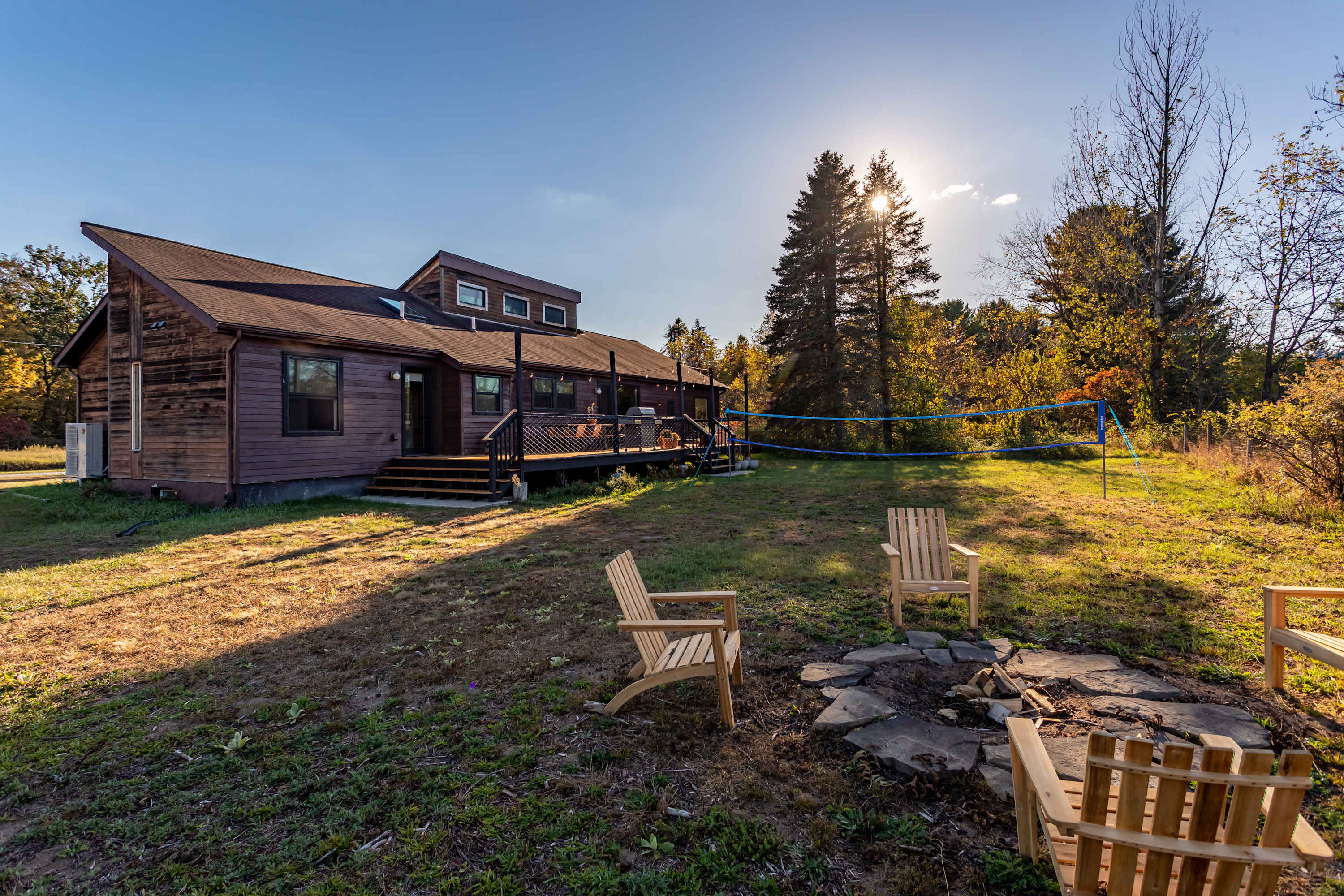 The back of the house features a large wooden deck with outdoor seating, overlooking a spacious grassy yard with a fire pit surrounded by Adirondack chairs. A volleyball net is set up, and mature trees line the property.