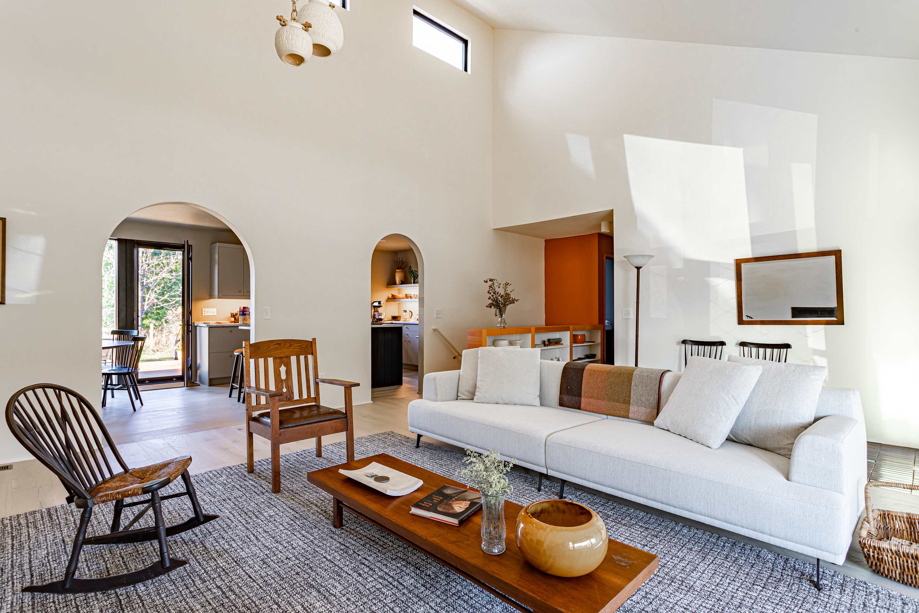 A spacious living room boasts high ceilings and light wood floors, furnished with a long white sofa adorned with a colorful throw, a wooden coffee table, and a rocking chair. A large rug defines the seating area, with arched doorways visible in the background.