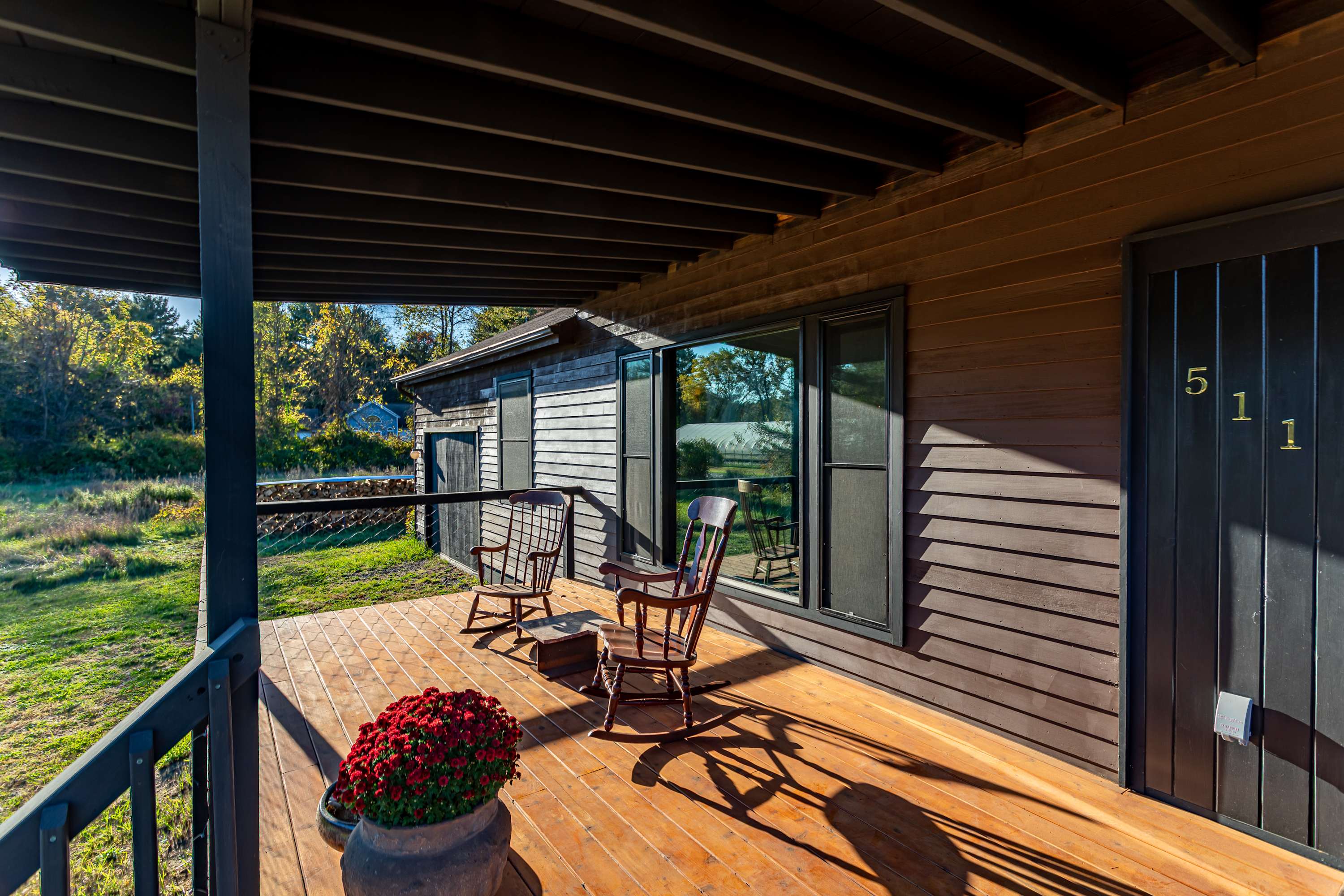 A covered wooden porch with two rocking chairs and potted red mums, offering a cozy outdoor seating area with views of the surrounding grassy yard and trees.