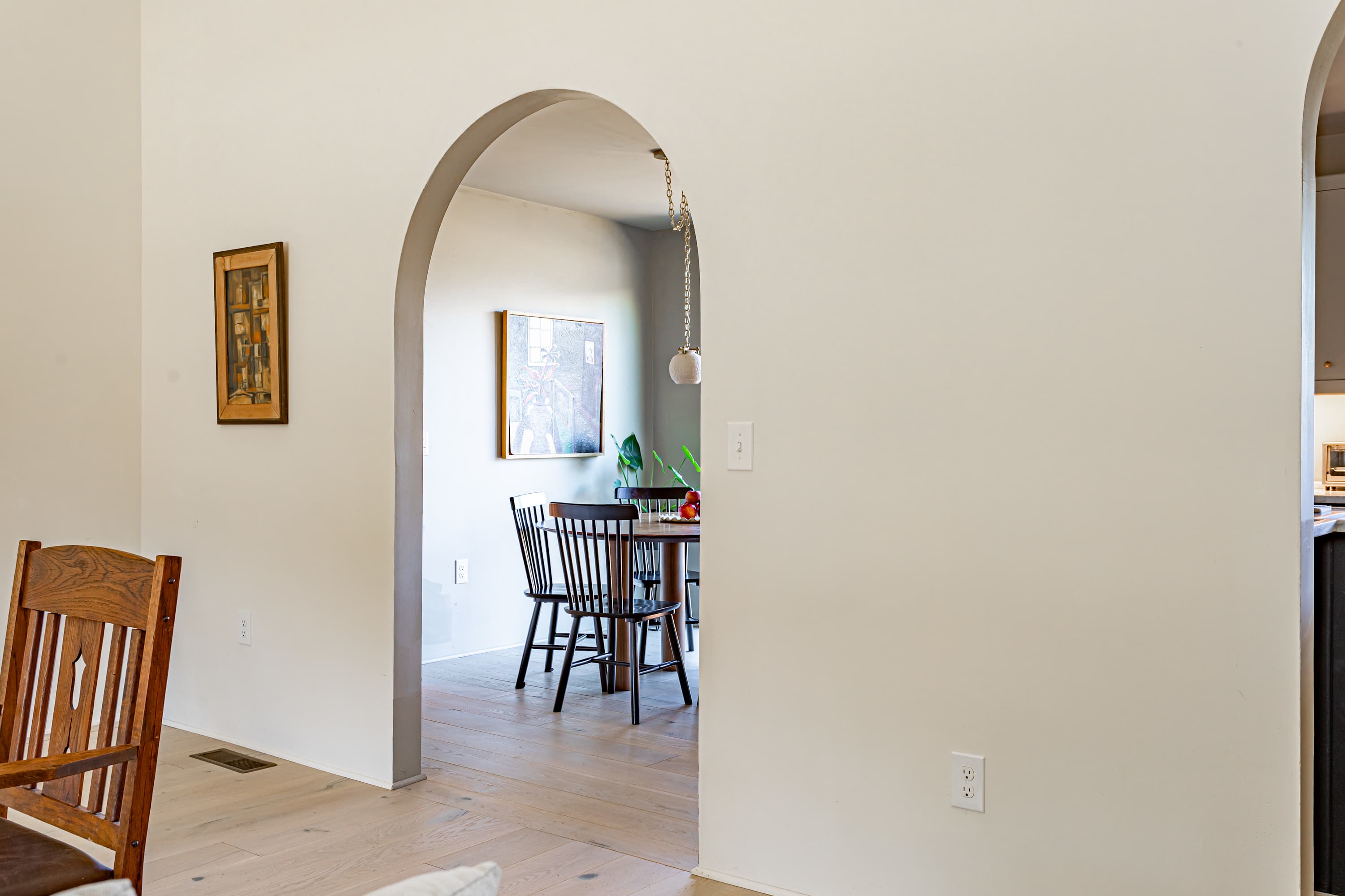 A view through a graceful archway from the living area into the dining room, revealing a round wooden table and black chairs, with light hardwood floors throughout.