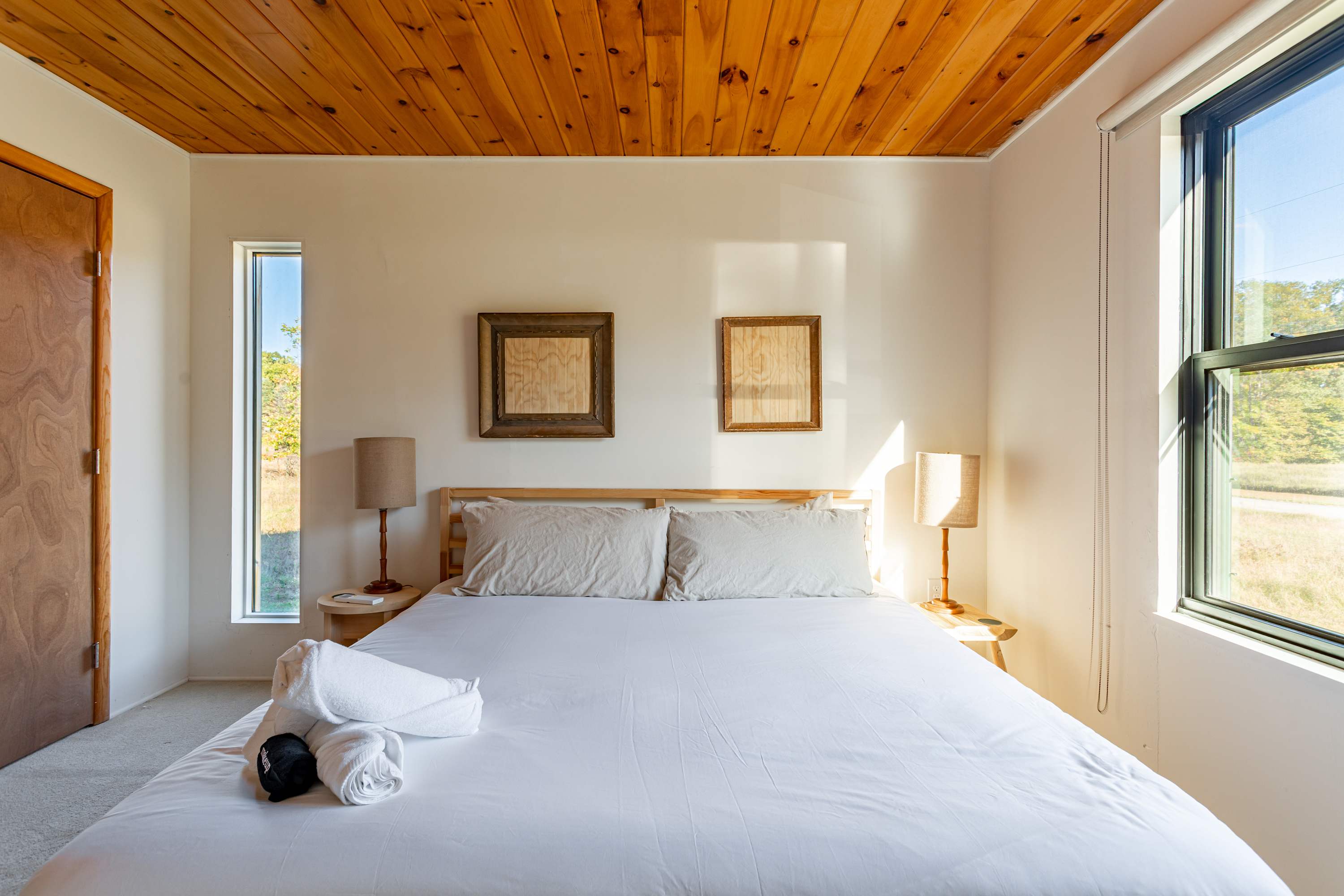 Serene bedroom featuring a queen-sized bed with white linens, flanked by two wooden nightstands with lamps, under a warm wooden plank ceiling. Two framed artworks adorn the wall, and large windows offer natural light.