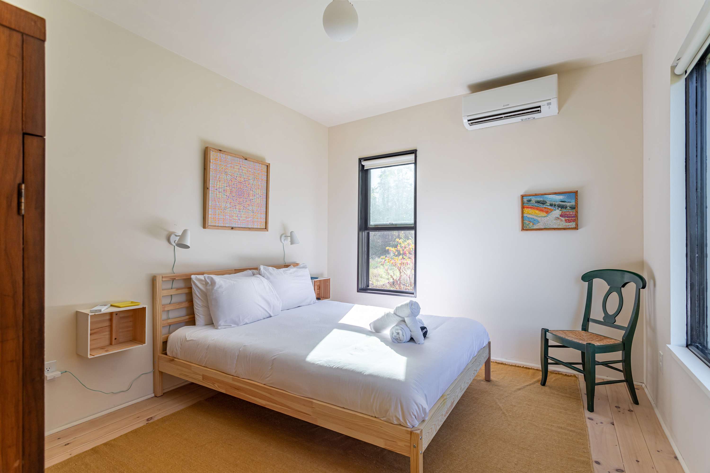 A bright bedroom with a queen-sized bed featuring a light wood frame and white bedding, flanked by floating wooden nightstands. A large window offers natural light, complemented by a green wooden chair and an AC unit.