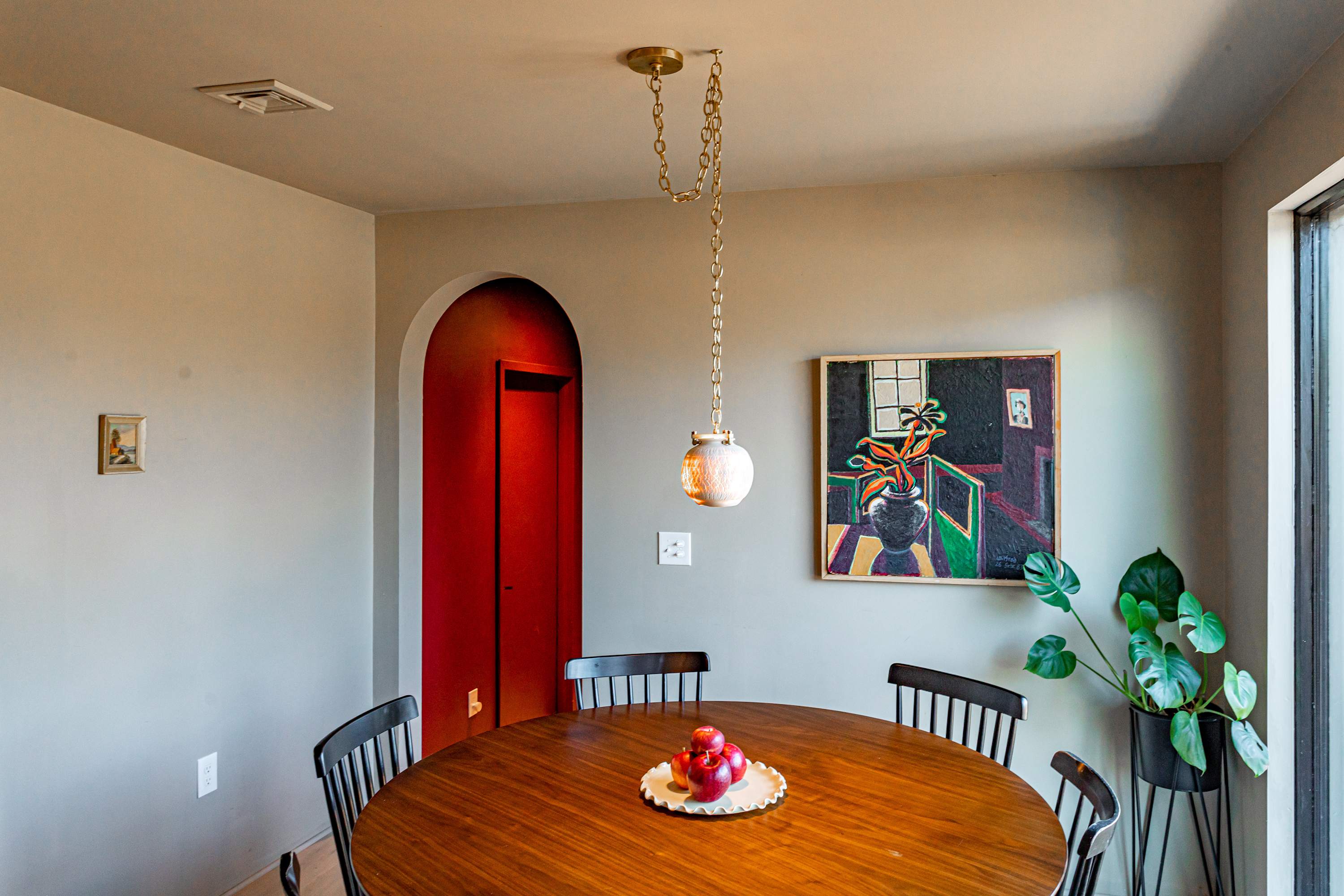 Inviting dining area with a round wooden table and black chairs, illuminated by a unique pendant light. A colorful abstract painting hangs on the light grey wall, and an arched red doorway adds a pop of color.