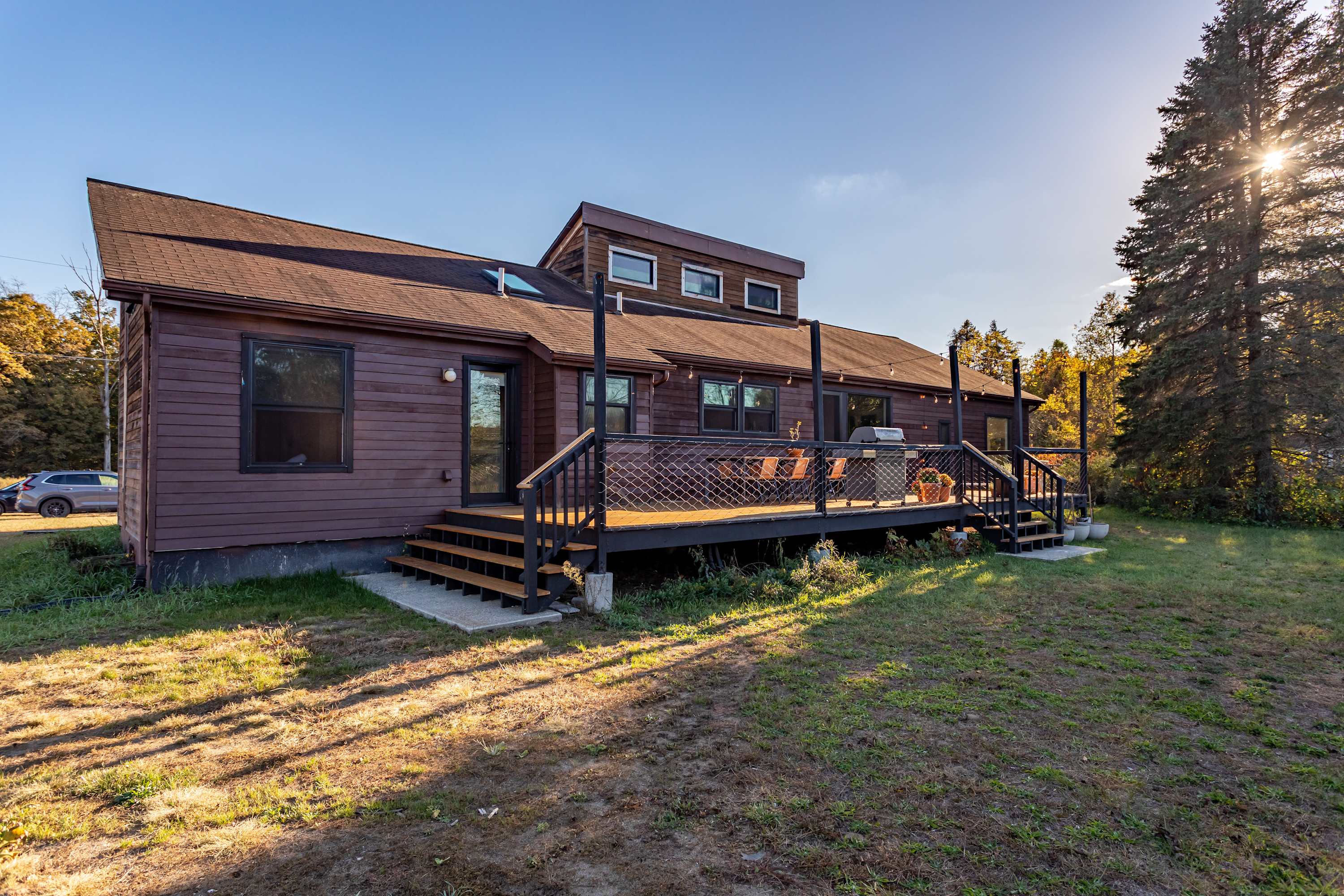 The back of the house features dark brown siding and a large wooden deck with black railings and mesh infill. Outdoor furniture is visible on the deck, surrounded by a grassy yard and mature trees under a clear sky.