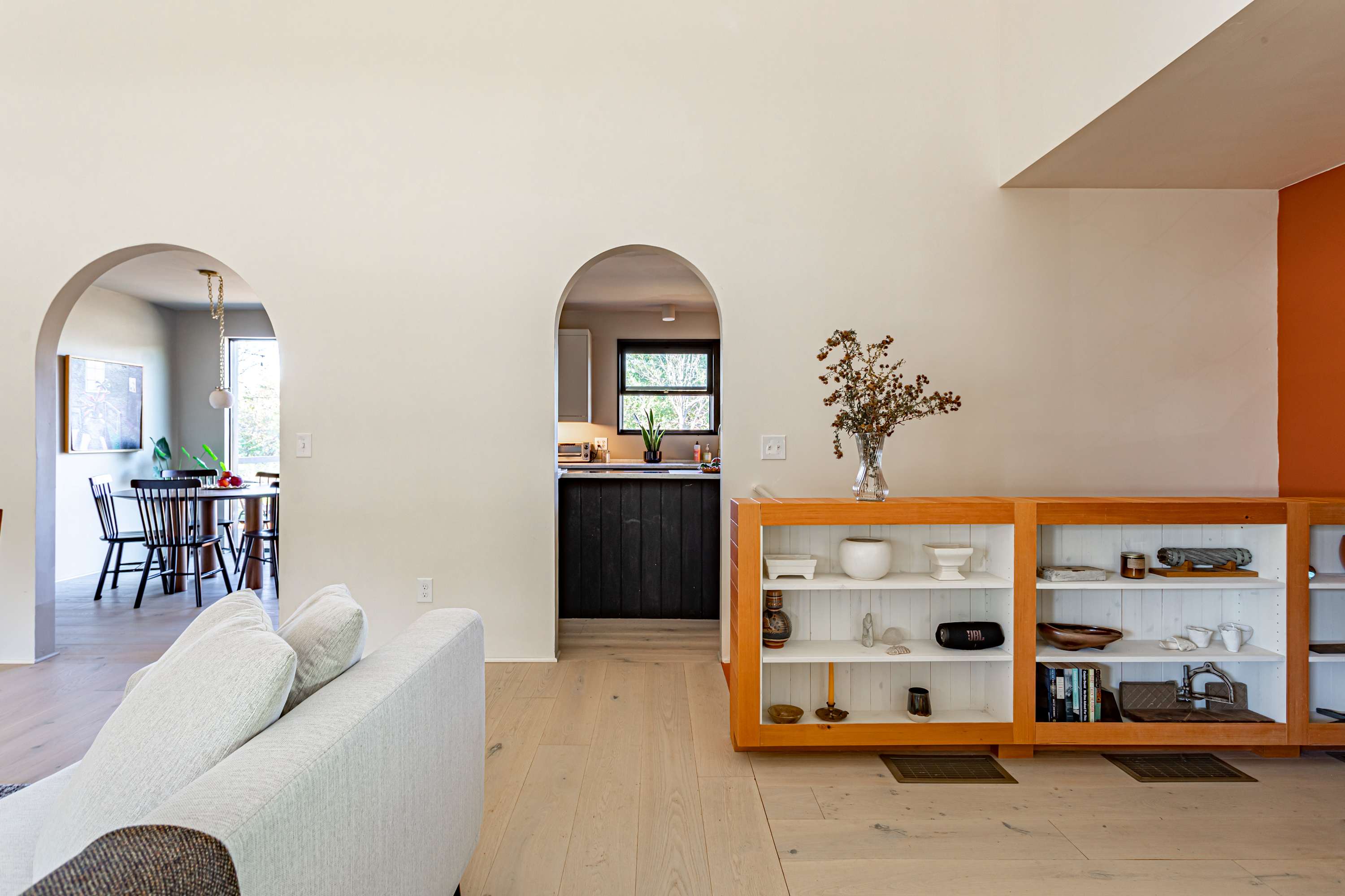 A bright and airy living room features light wood floors and white walls, with two arched doorways leading to the dining area and kitchen. A long wooden bookshelf filled with decorative items sits against the wall, complementing the modern aesthetic.