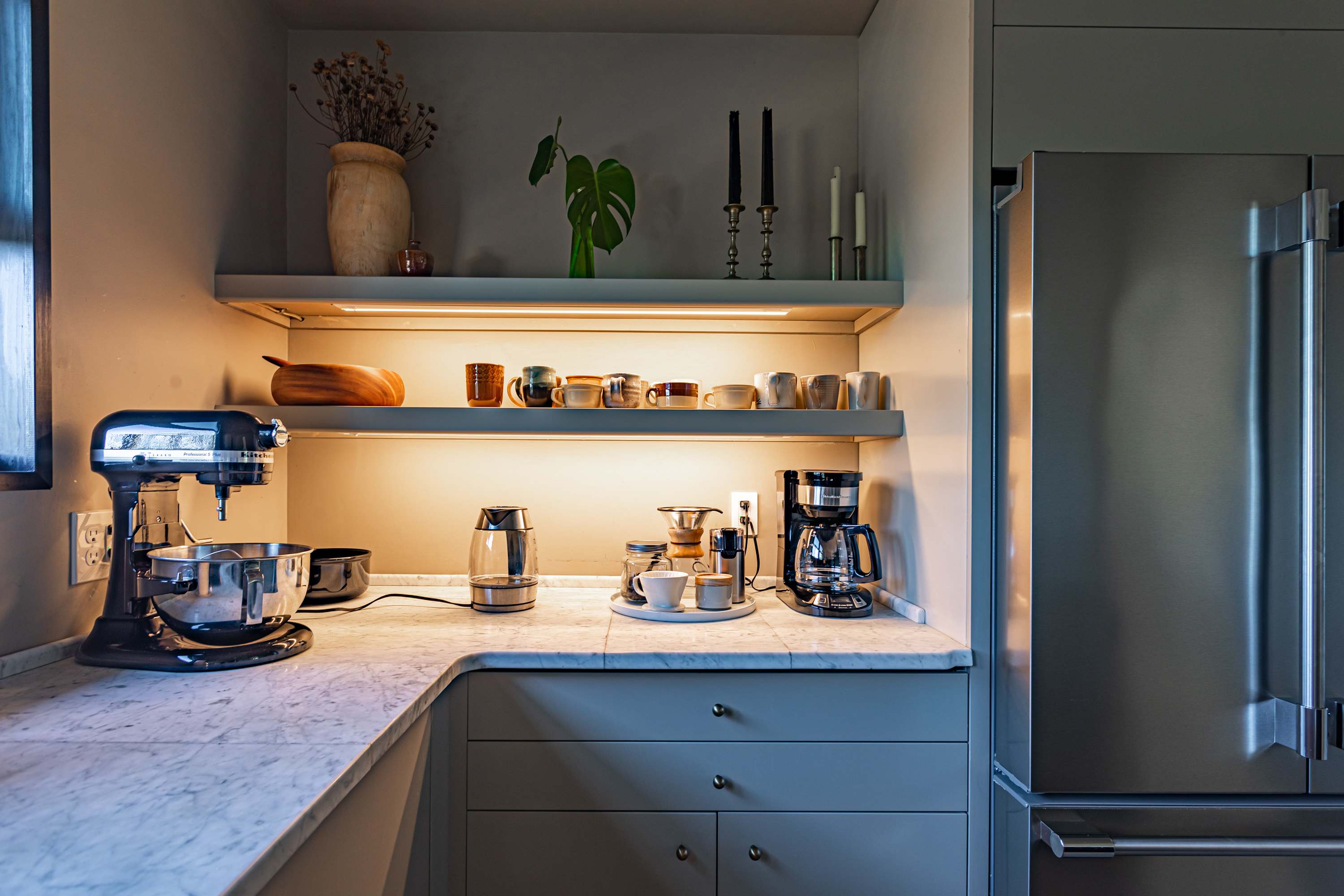 A well-equipped kitchen features light grey cabinetry, white marble countertops, and open shelving displaying various mugs and kitchenware. Modern appliances like a stand mixer, kettle, and coffee makers are neatly arranged on the counter.