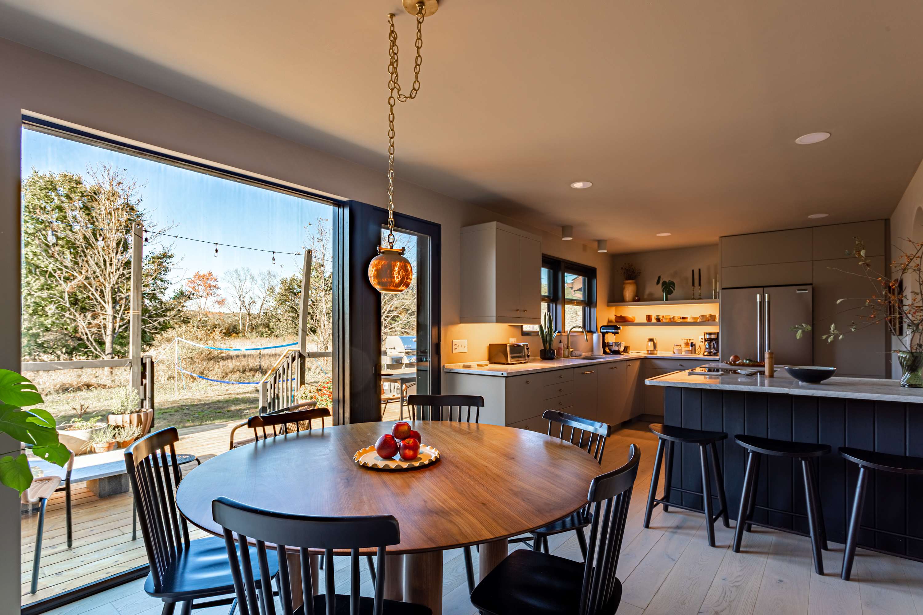 Bright dining area with a round wooden table and black chairs, adjacent to a modern kitchen and large glass doors opening to an outdoor deck. The kitchen features light grey cabinets and marble countertops.