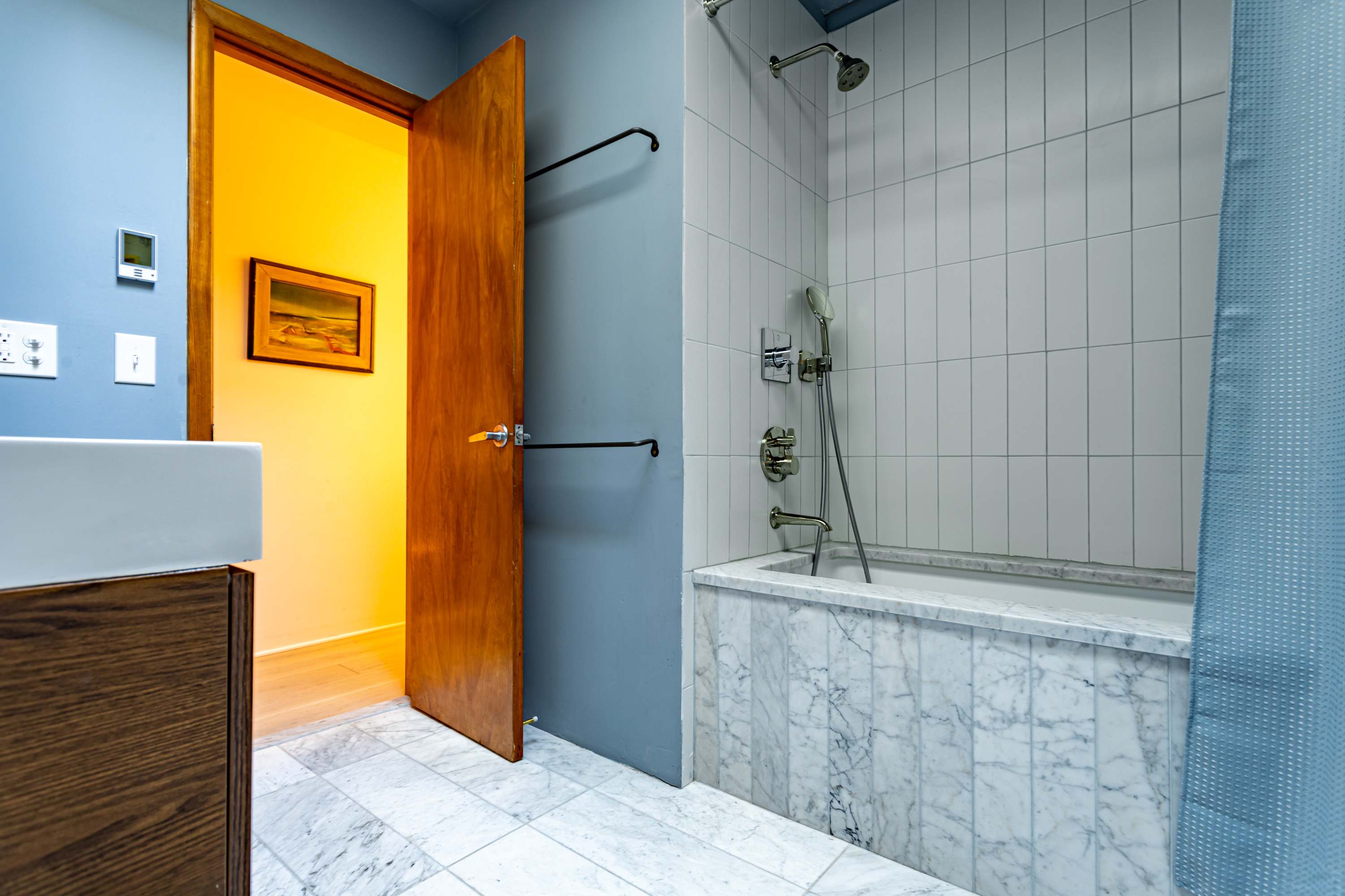 A close-up view of the white bathtub with marble tile surround and white subway tiles, featuring a modern shower head and faucet. A wooden door is open, revealing a warm-toned hallway.