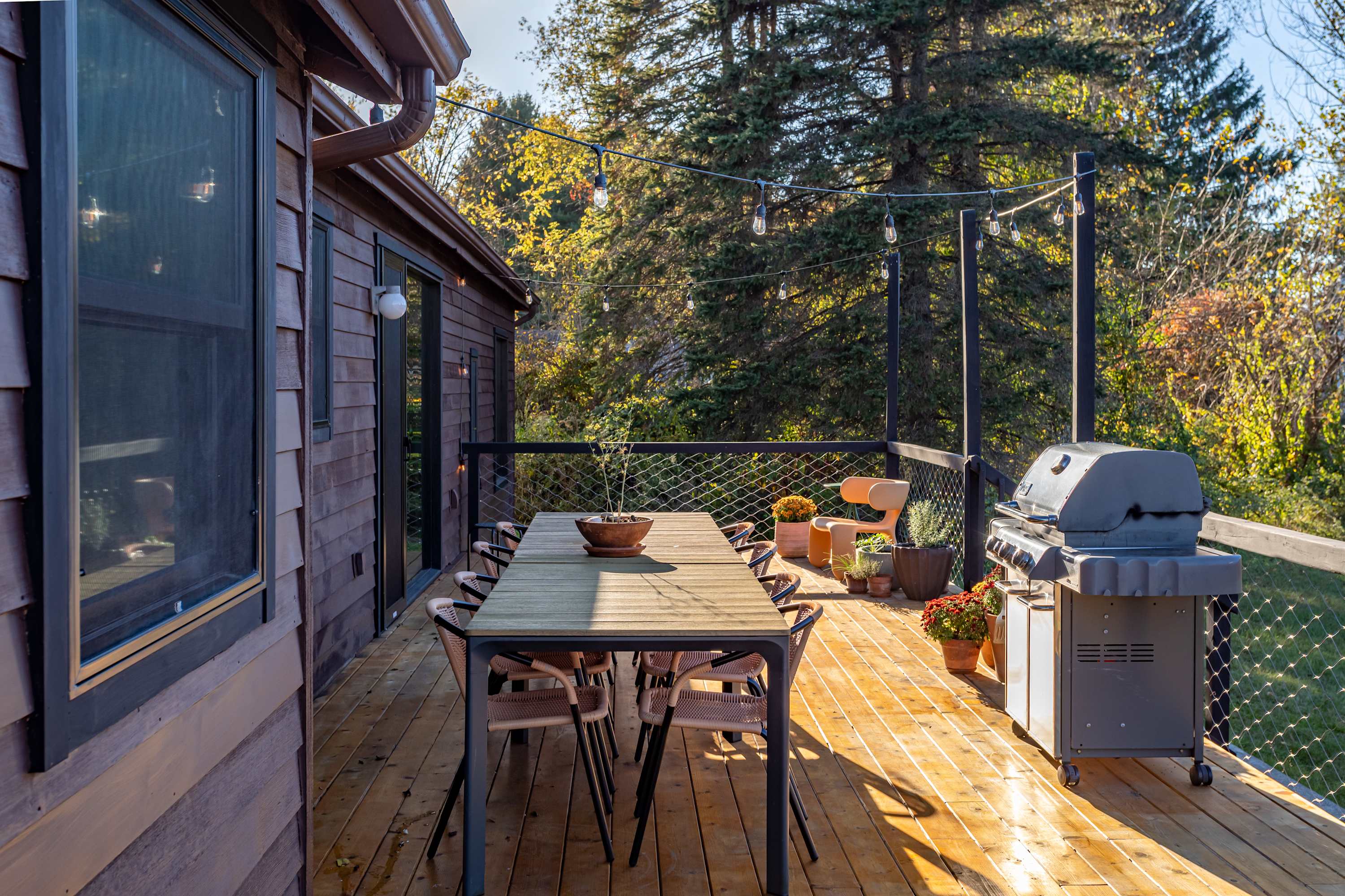 A spacious outdoor deck with a long dining table and chairs, a BBQ grill, and potted plants, surrounded by lush trees and strung with festive lights, perfect for outdoor entertaining.