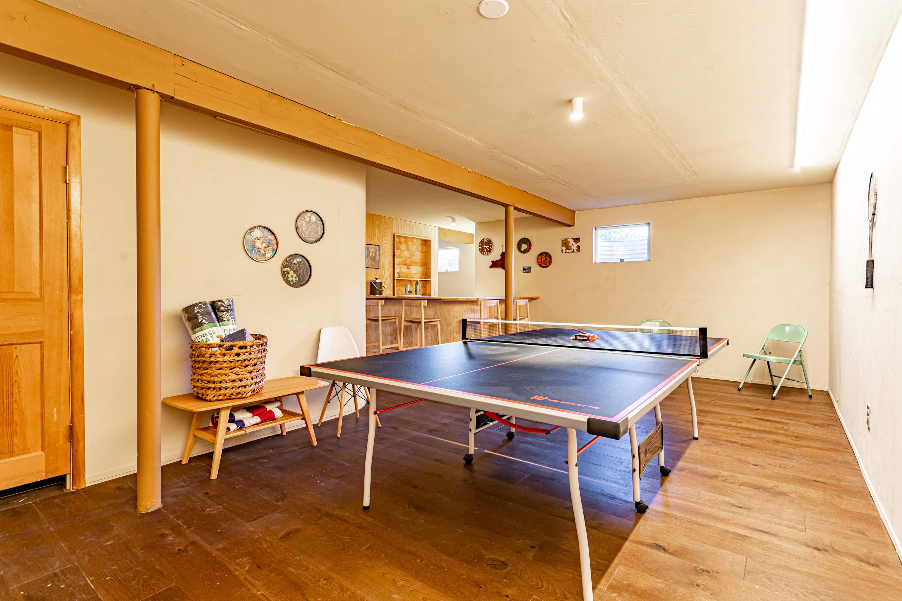 A spacious game room featuring a black and red ping pong table as the centerpiece, set on dark wooden floors. Exposed wooden beams and a distant bar area contribute to a recreational atmosphere.