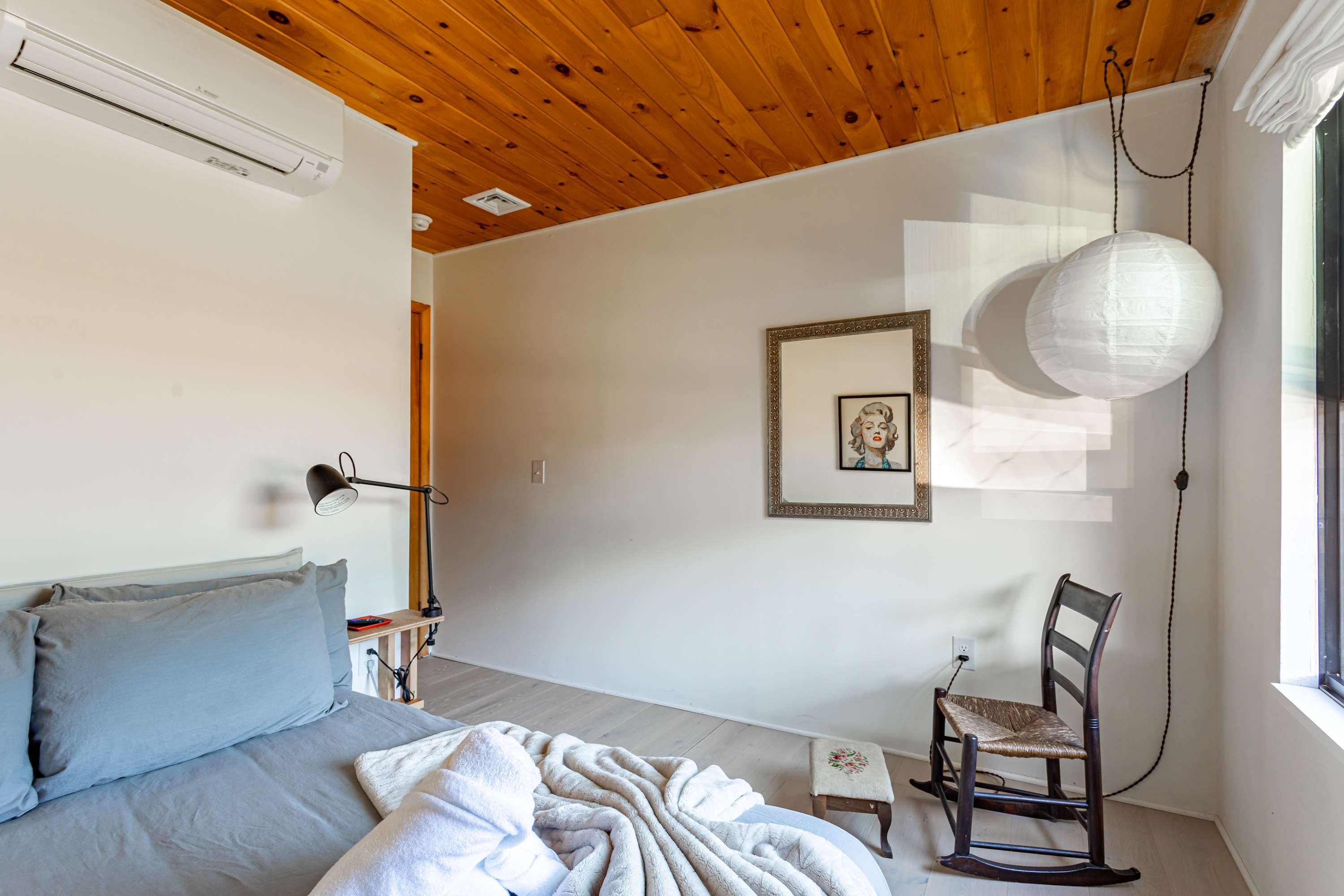 A bright bedroom with a single bed, light hardwood floors, and a warm wooden plank ceiling. A large mirror and a paper lantern add decorative touches, with a rocking chair in the corner.