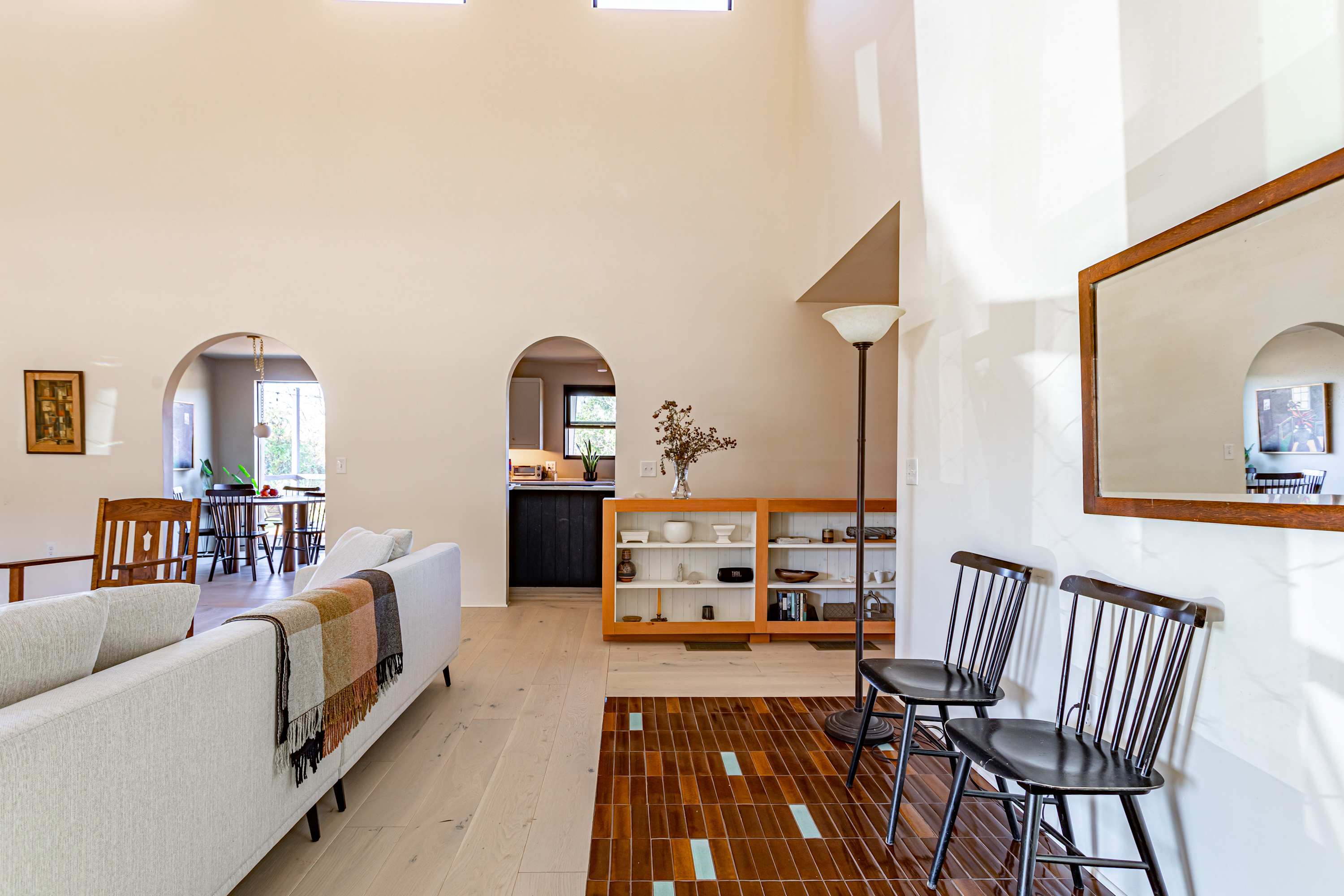 Bright, open-concept living space with light wood floors, a white sofa, and arched doorways leading to a dining area and kitchen. A wooden bookshelf with decor and two black chairs sit on a tiled rug.