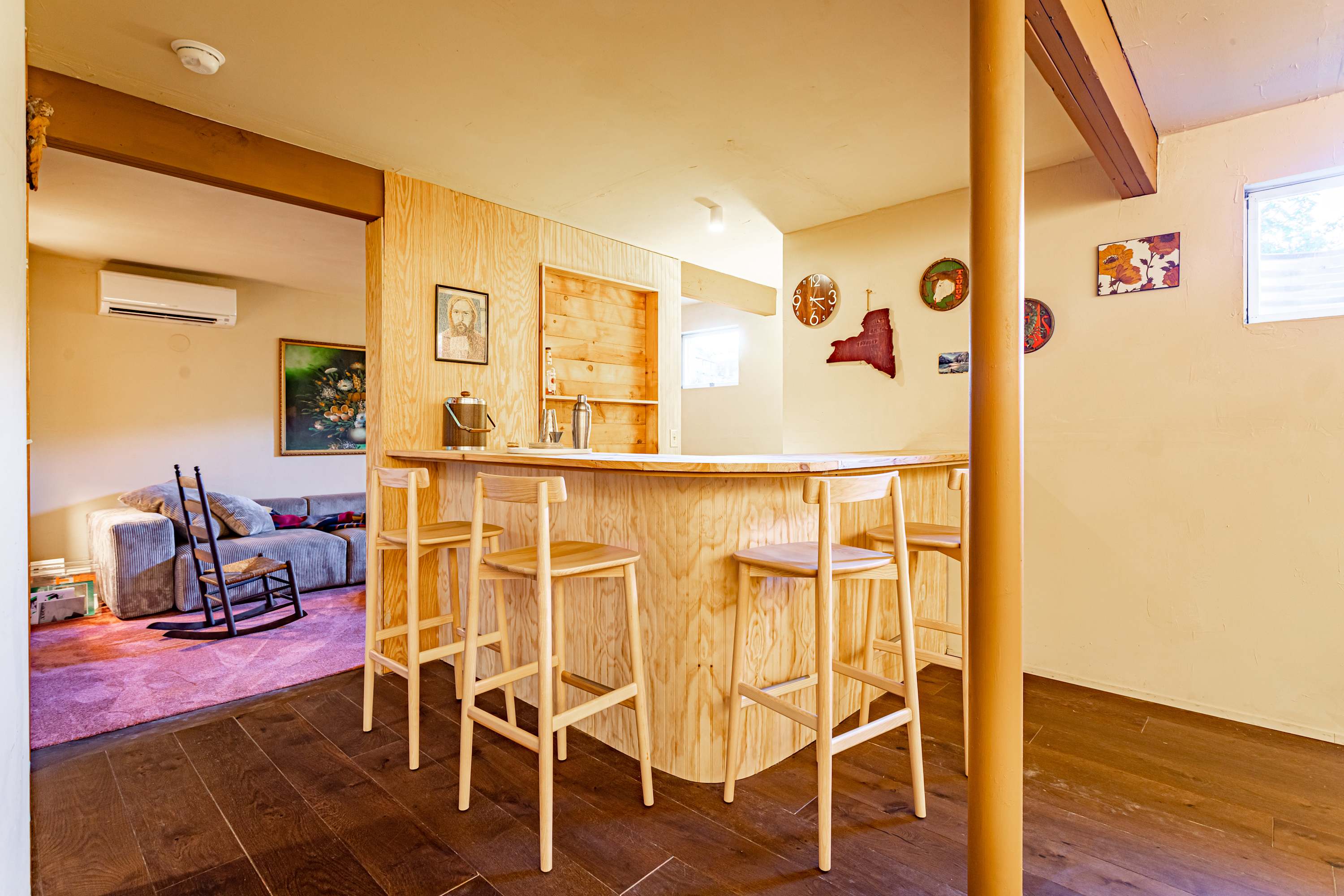 A cozy basement bar area features a light wood curved bar with four matching high-back stools, set against a plywood-paneled wall adorned with decorative items. In the background, a living area with a grey sofa and rocking chair is visible.