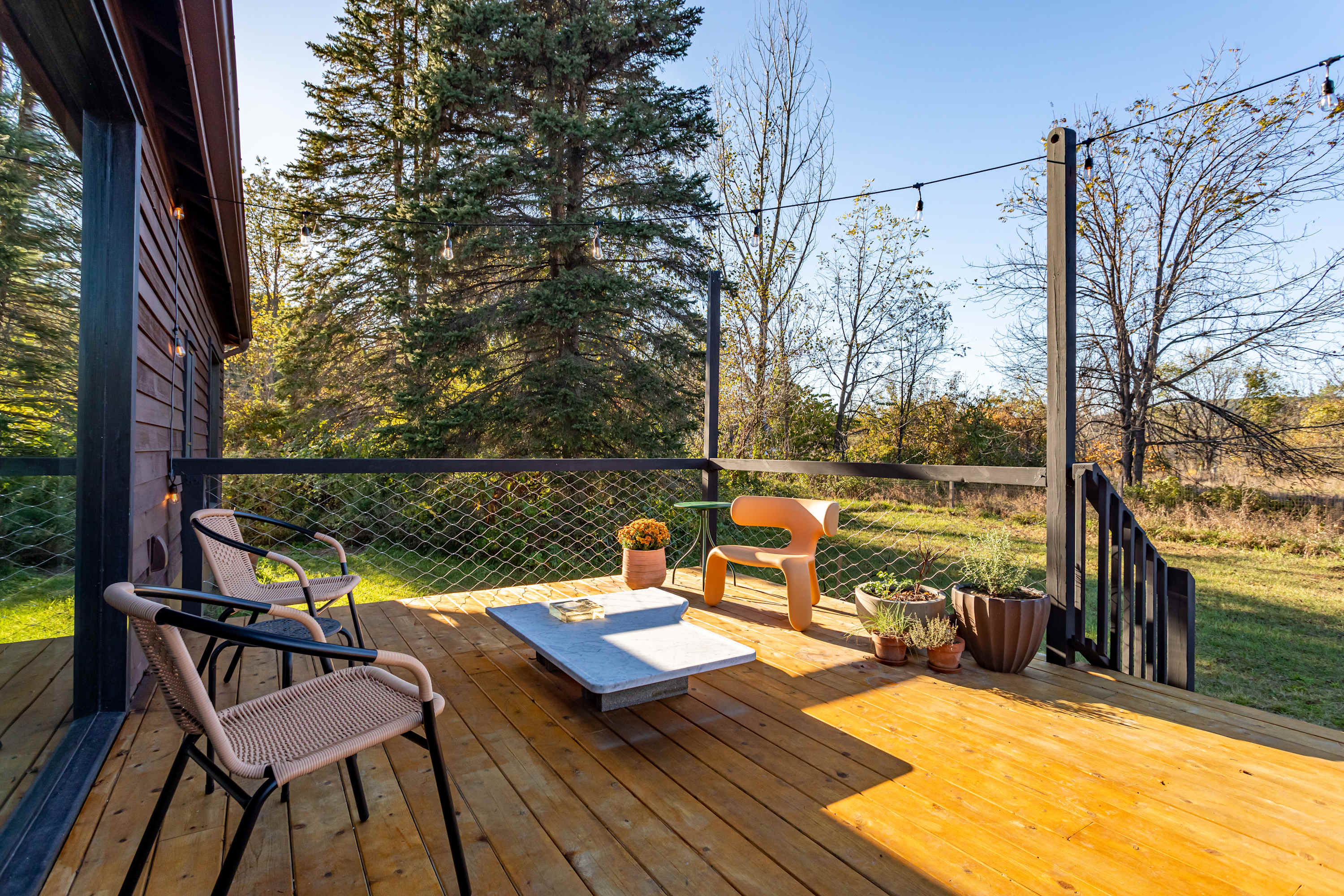 A sunny wooden deck offers a relaxed seating area with a modern concrete coffee table, two woven-back chairs, and a unique orange lounge chair. The area is enclosed by a black railing with wire mesh, overlooking a grassy backyard and trees.
