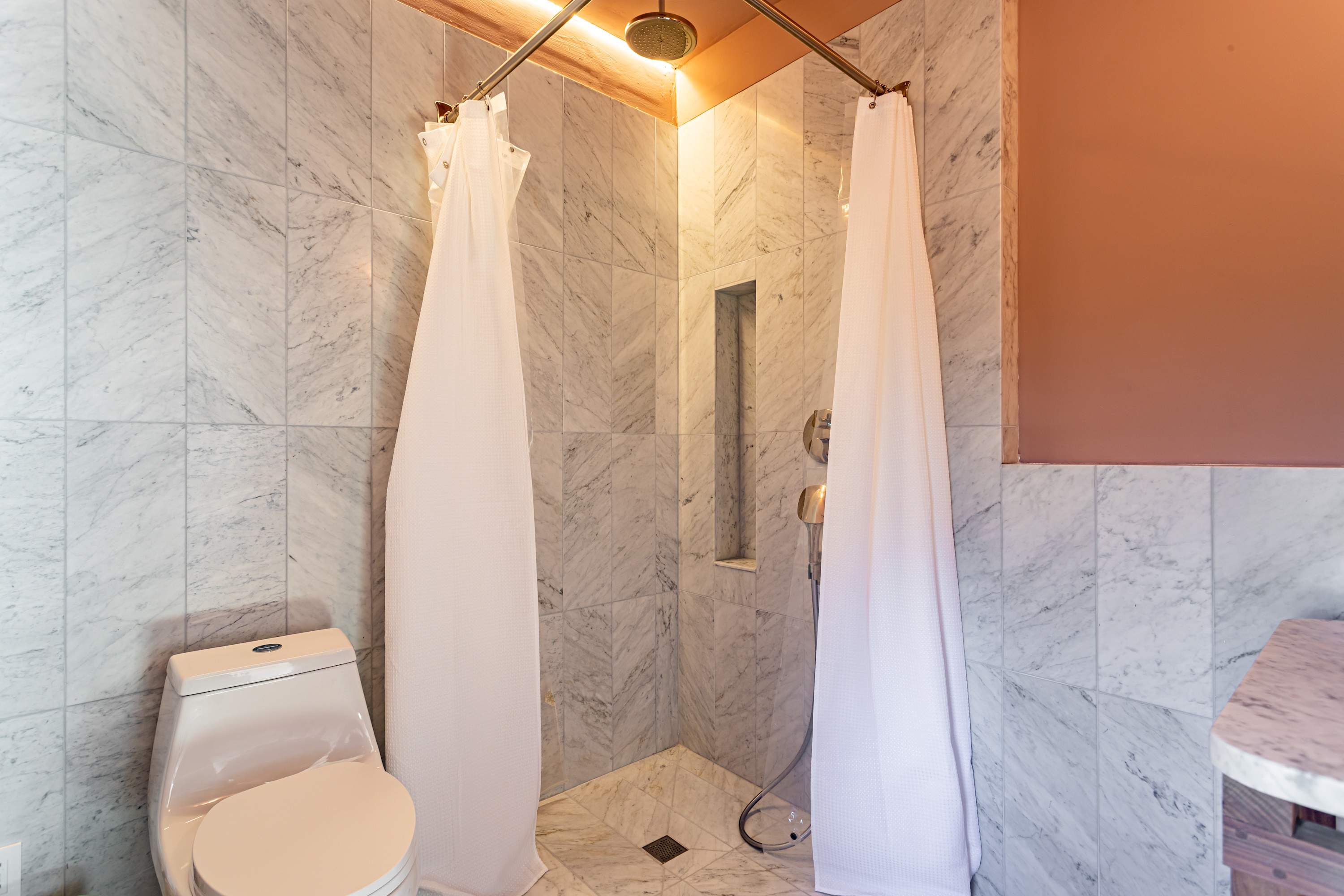 Elegant shower area with white marble tile walls and floor, featuring a corner shower with two white curtains and a rain shower head. A white toilet is visible next to the shower.