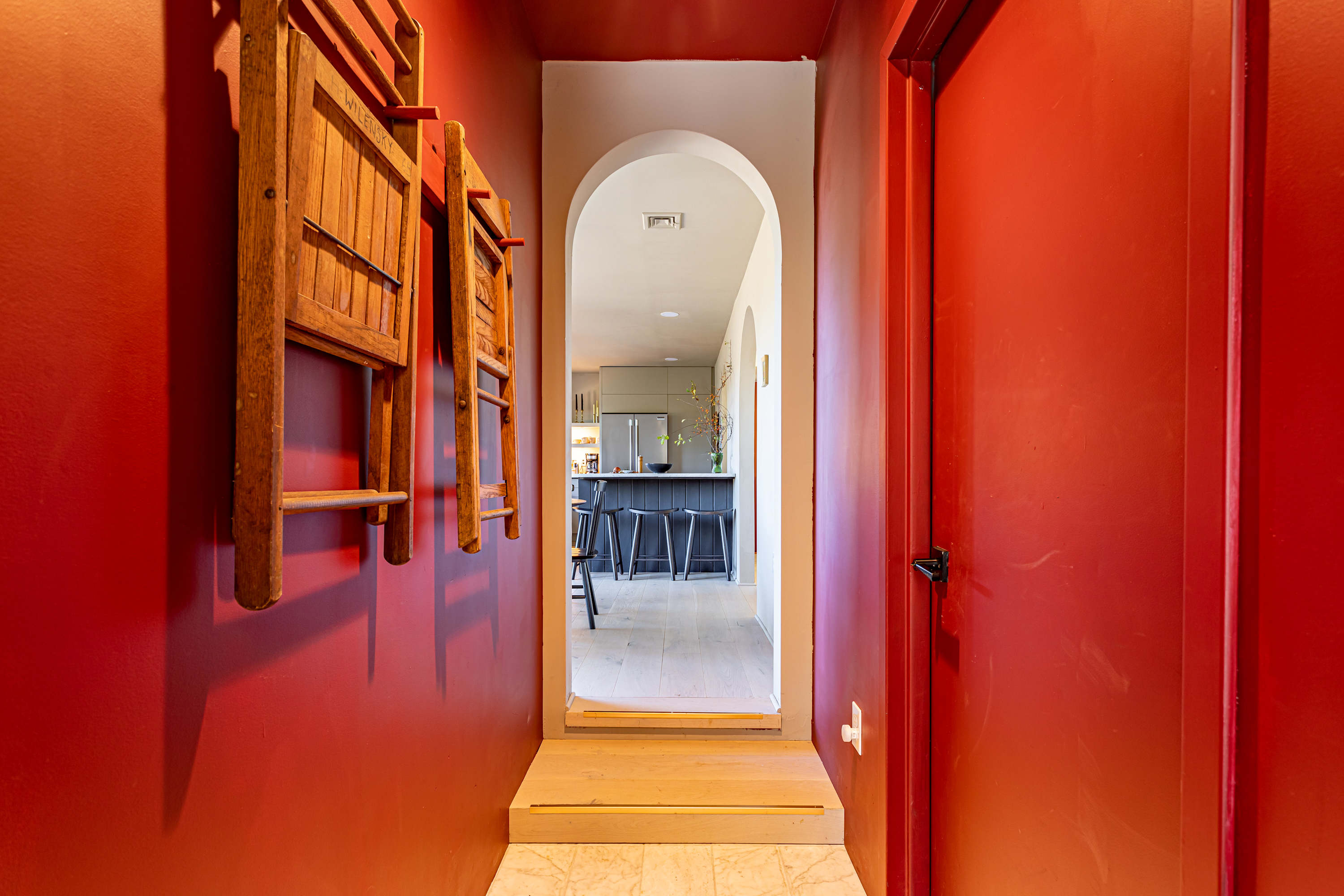 Striking red hallway with a wooden folding chair mounted on the wall, leading to an arched doorway that opens into a bright kitchen area. The floor transitions from light tile to light wood.