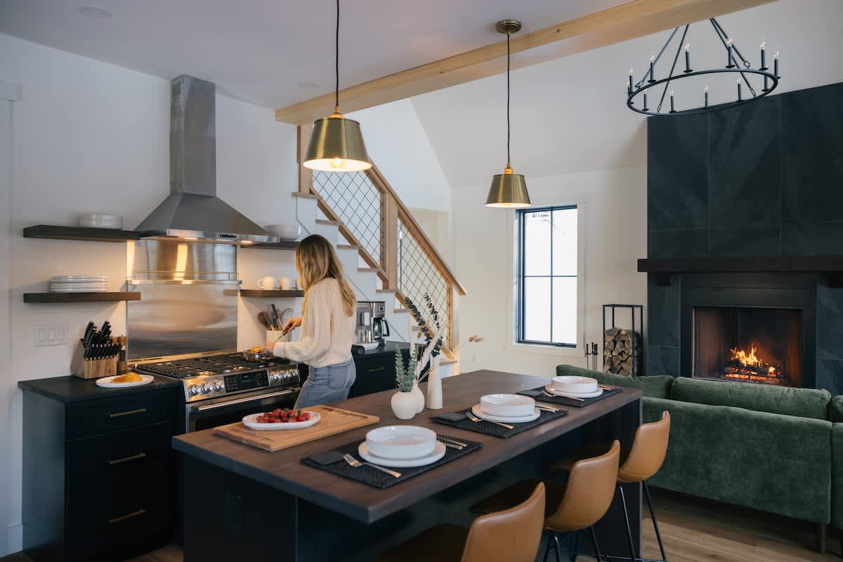 A woman cooks at a modern kitchen island with dark cabinetry and brass pendant lights, overlooking a living area with a fireplace. The kitchen features stainless steel appliances and a large range hood.