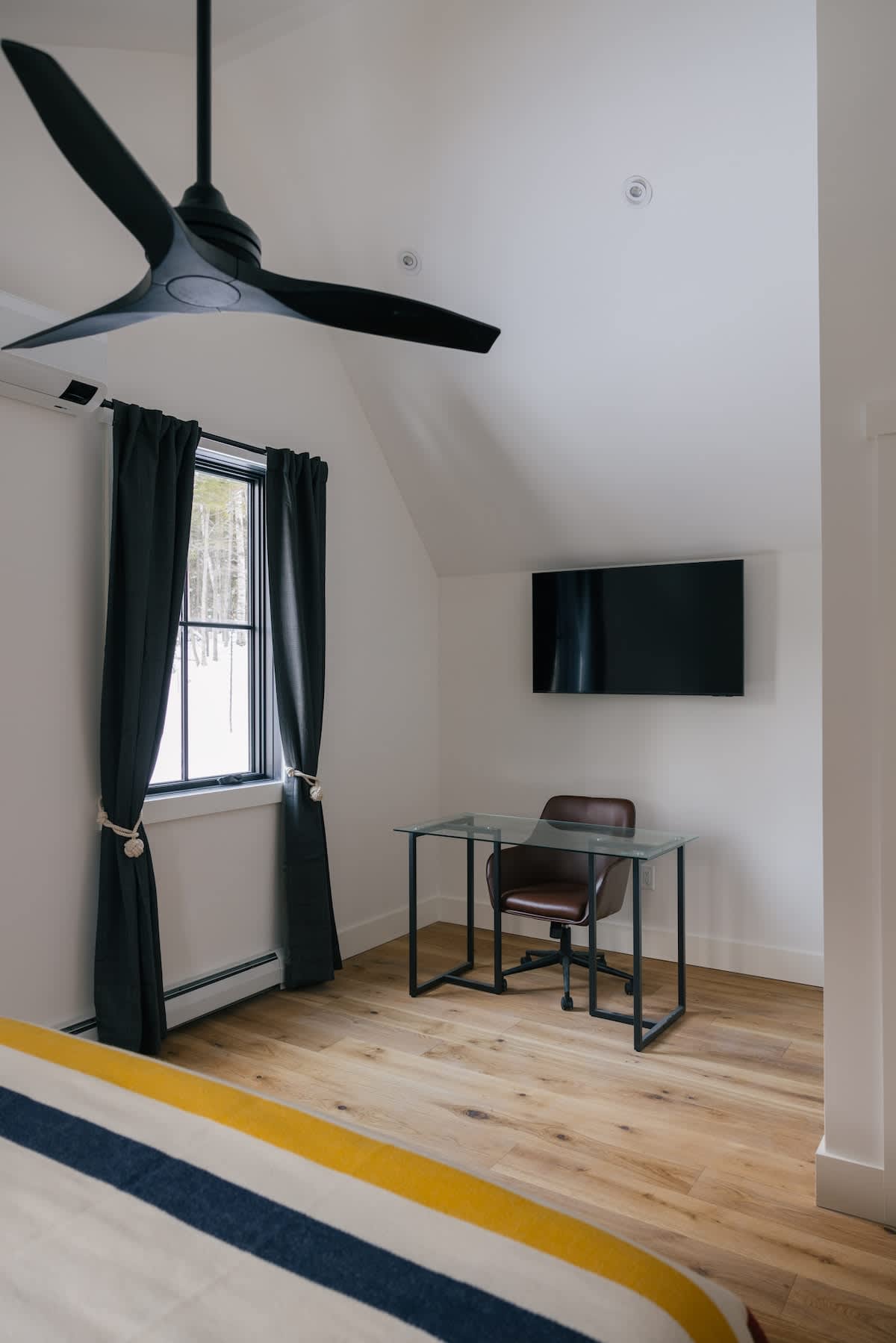 Bright bedroom with light wood floors, a glass-top desk, and a brown office chair. A wall-mounted TV and a modern black ceiling fan are visible, along with a window draped with dark curtains.