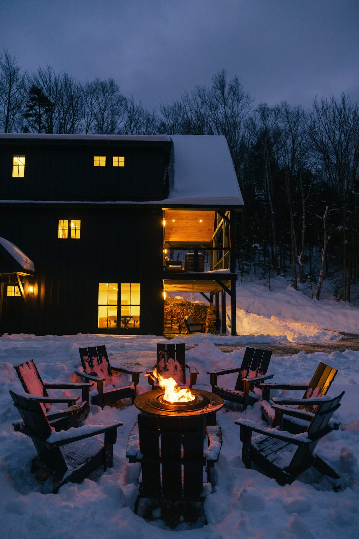 Evening view of a cozy outdoor fire pit surrounded by six dark Adirondack chairs, all covered in a light dusting of snow. The fire pit glows warmly, set against the backdrop of a dark modern house with illuminated windows and bare winter trees.