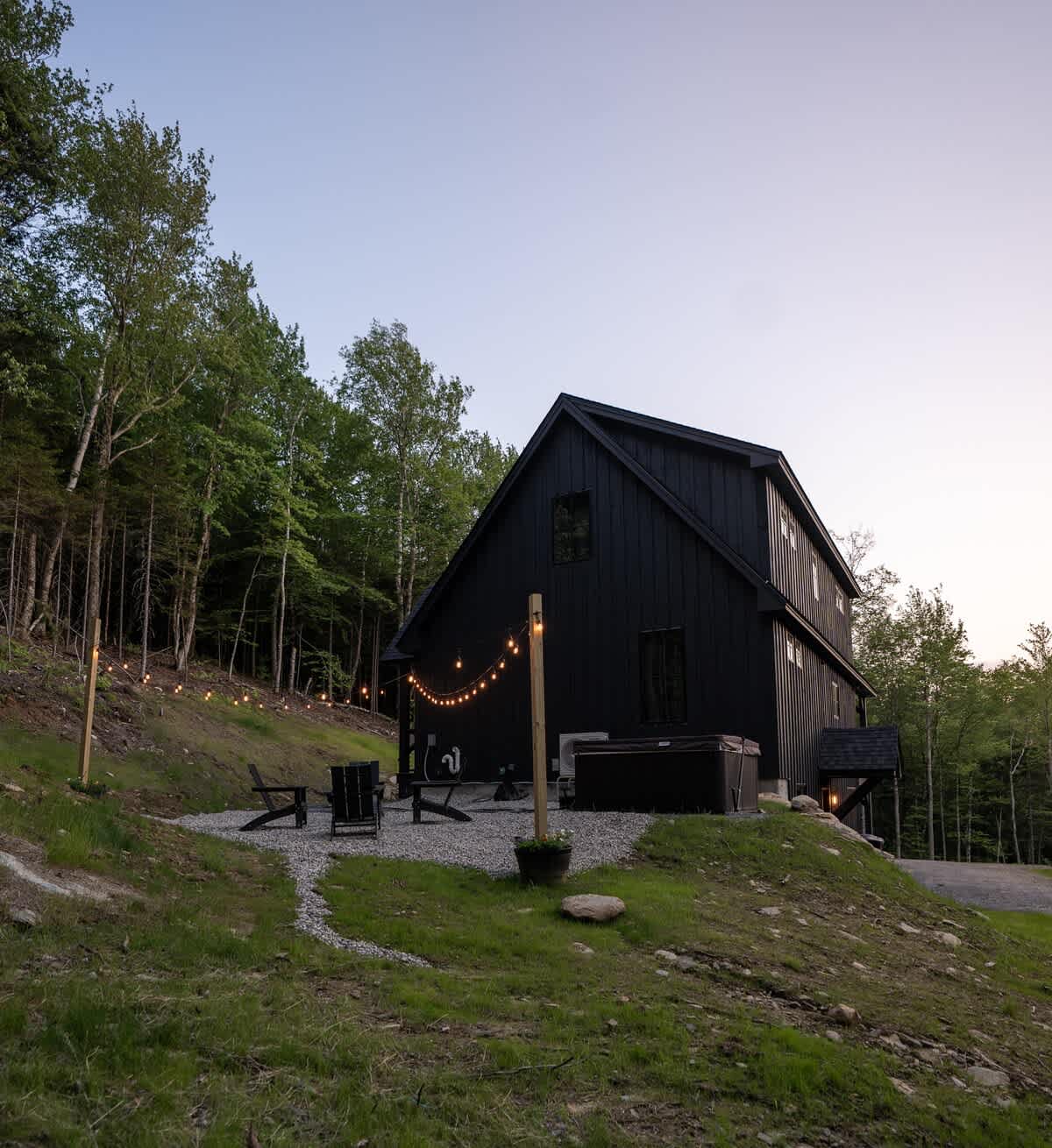 The modern black-sided cabin is viewed from the back, showcasing a gravel patio with a hot tub, Adirondack chairs, and string lights leading up a grassy hill. The lush green forest surrounds the property under a clear evening sky.
